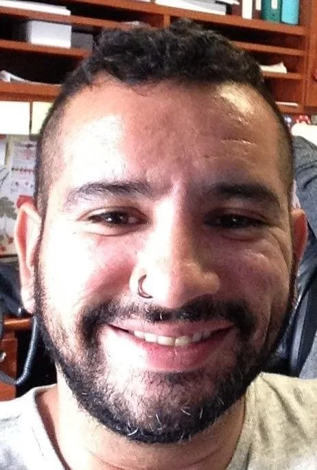 Close-up of a smiling man with a beard and mustache, wearing a nose ring, in an indoor setting with shelves and books in the background.
