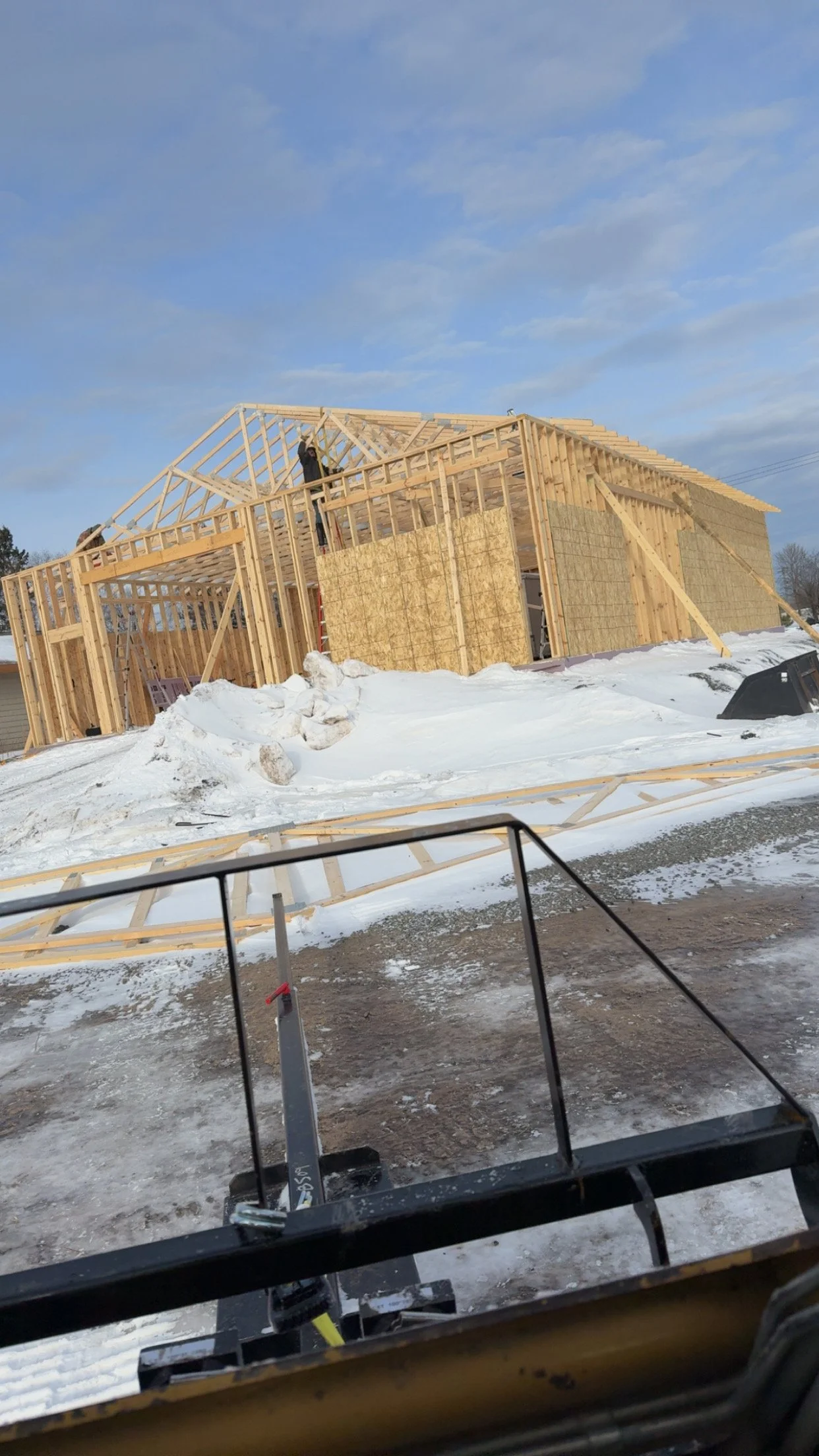 Construction of a house with wooden framing, snow on the ground, and a clear sky, with a construction worker working on the roof.