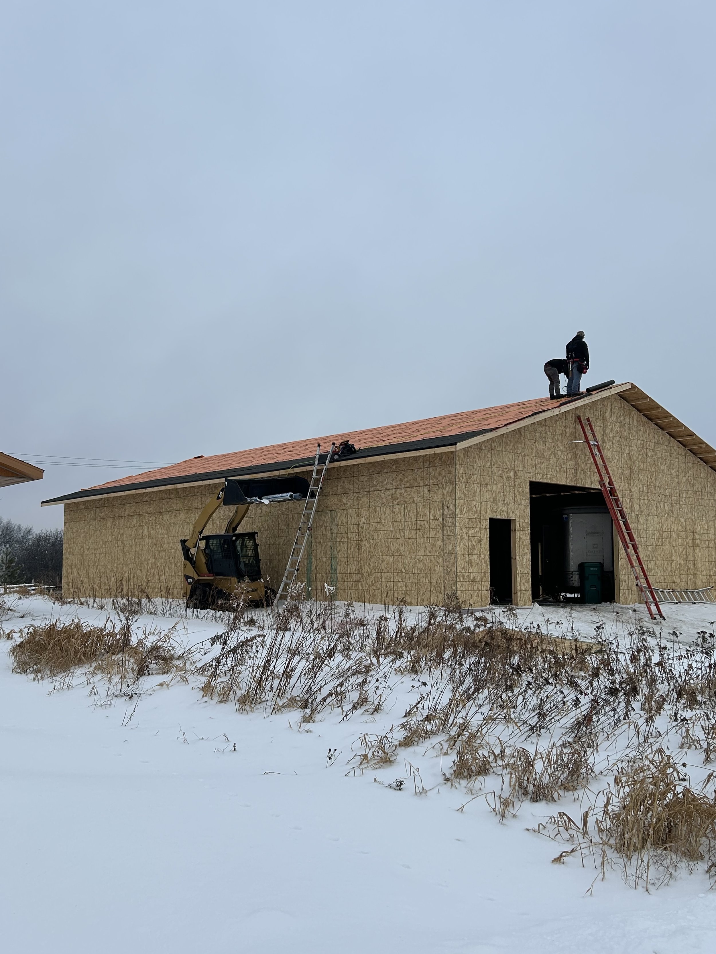 Construction workers on a roof of a house, with ladders leaning against the building, in a snowy landscape.