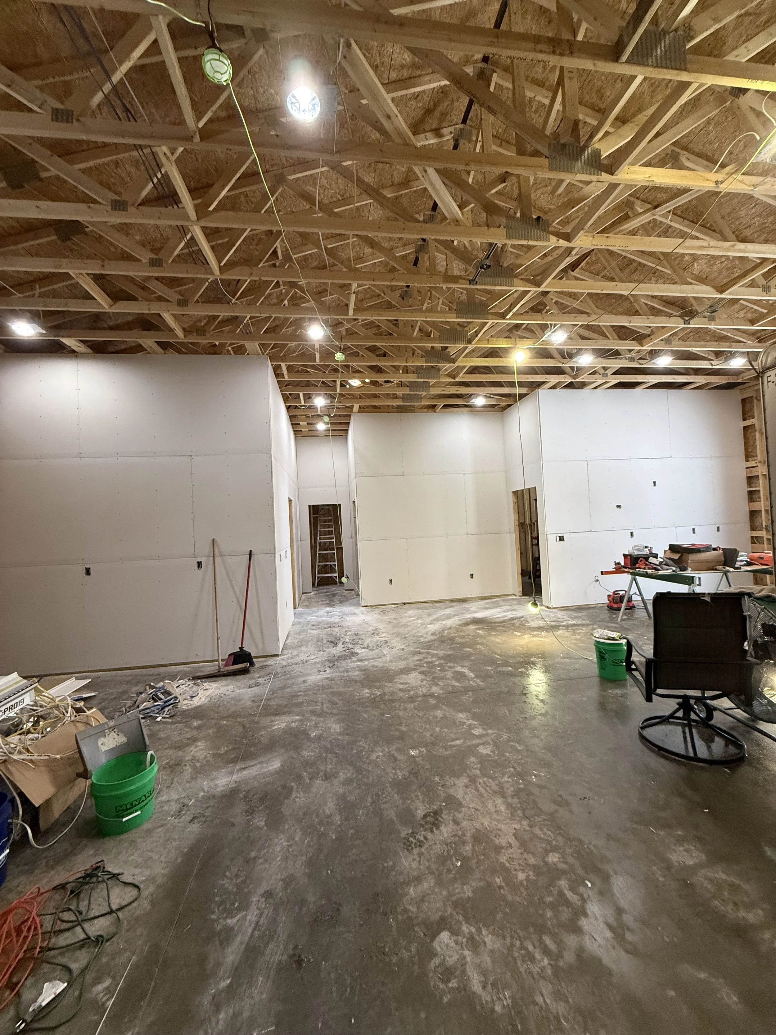 Interior view of a building under construction with unfinished drywall walls, exposed wooden ceiling trusses, and construction tools and materials scattered on the floor.