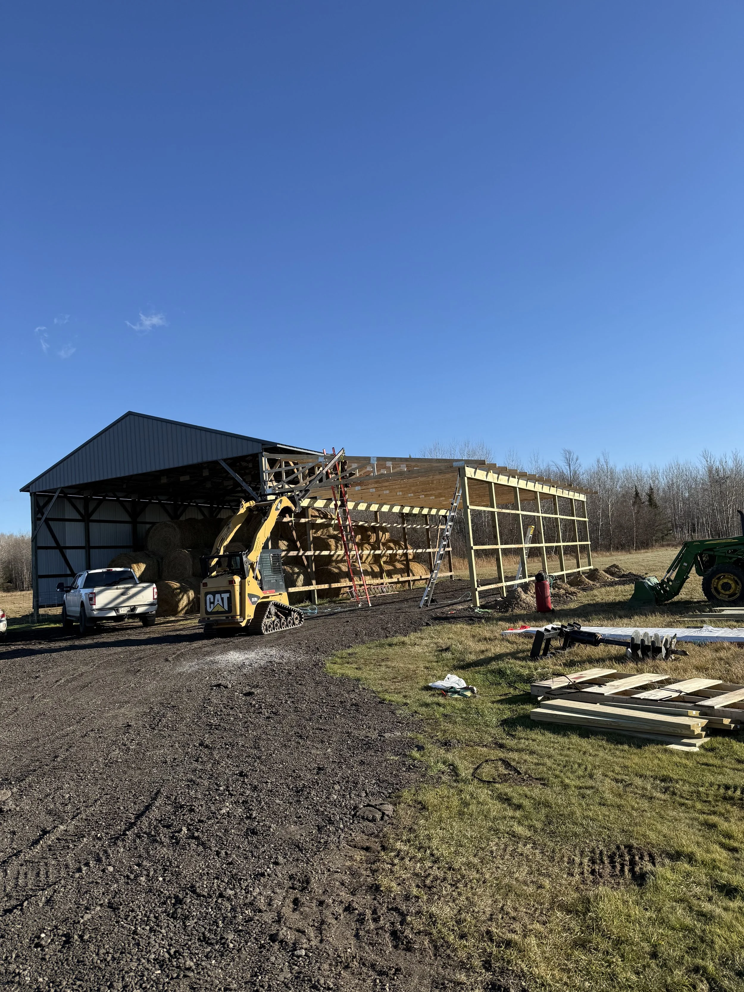 Construction site on a farm with a partially built wooden structure, a tractor, a pickup truck, and bales of hay under a clear blue sky.