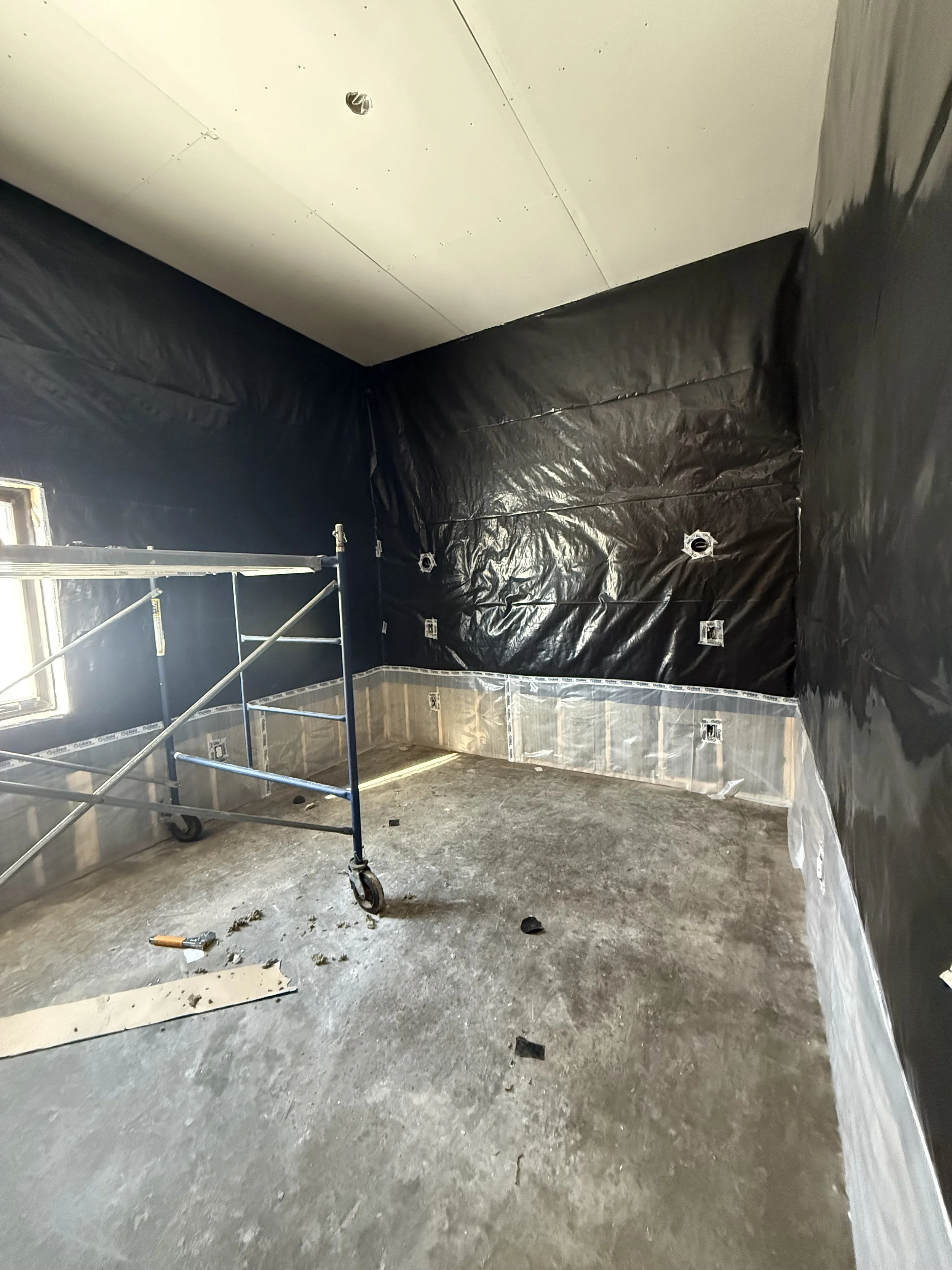 Room under construction with black plastic sheeting on the walls, electrical outlets, a small window, and a scaffold with wheels.