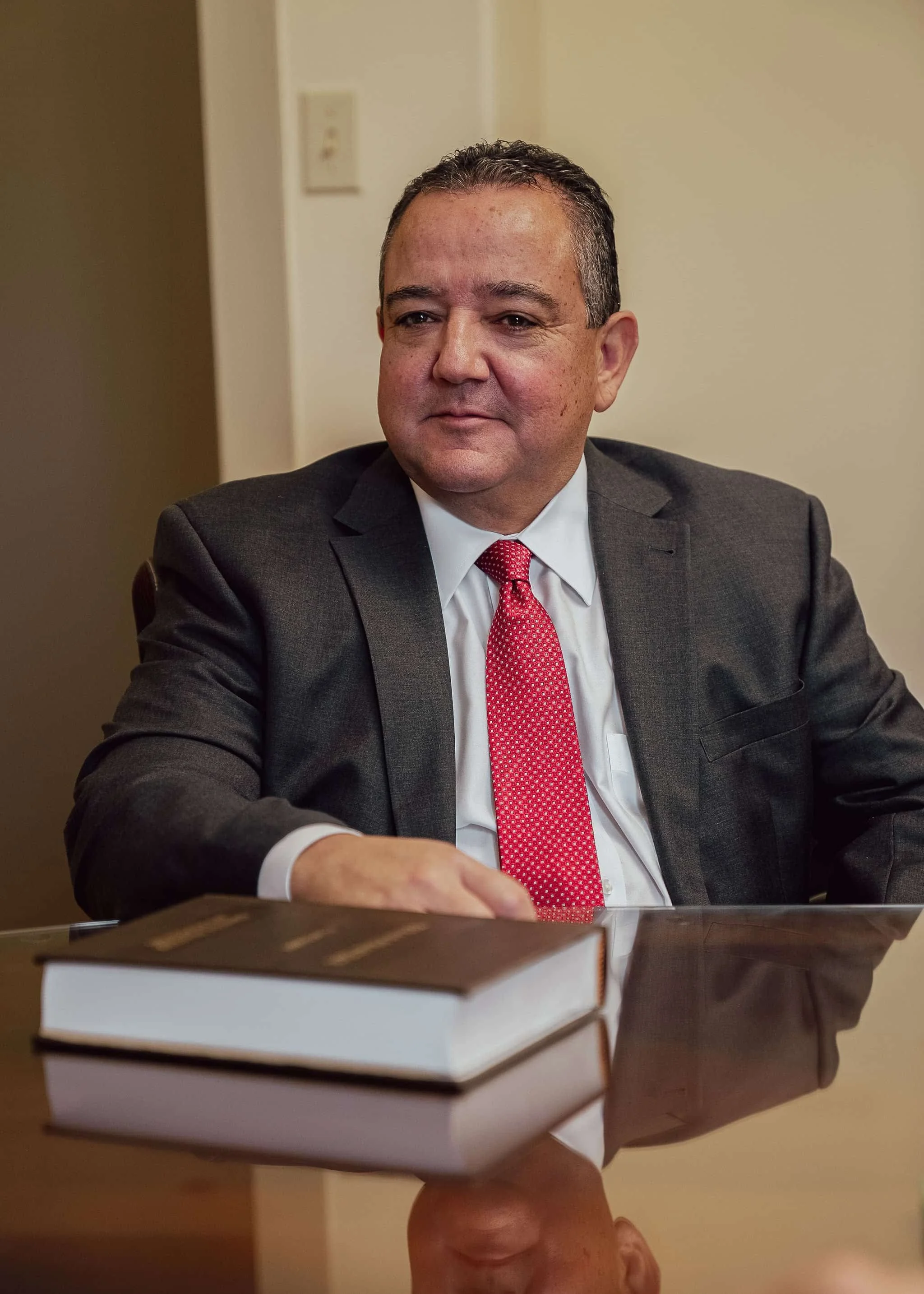 Fred Casillas, in a suit and red tie, sits at a desk with a book in front of him, gazing slightly to the side.