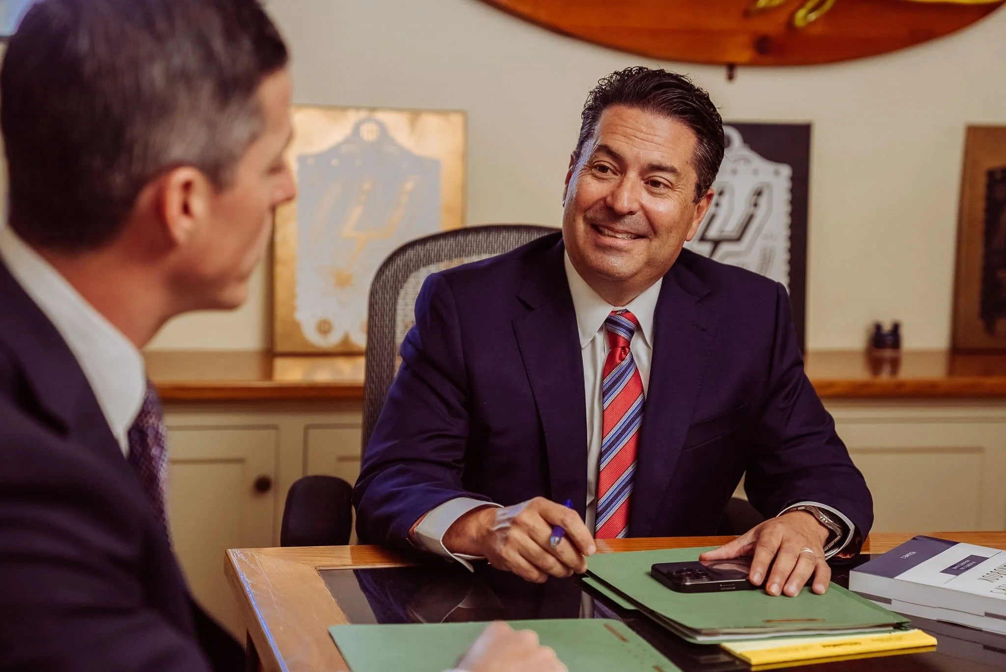 David Christian attorney and Thom Nisbet  in suits sit at a desk, smiling and talking during a meeting, with folders and documents in front of them.