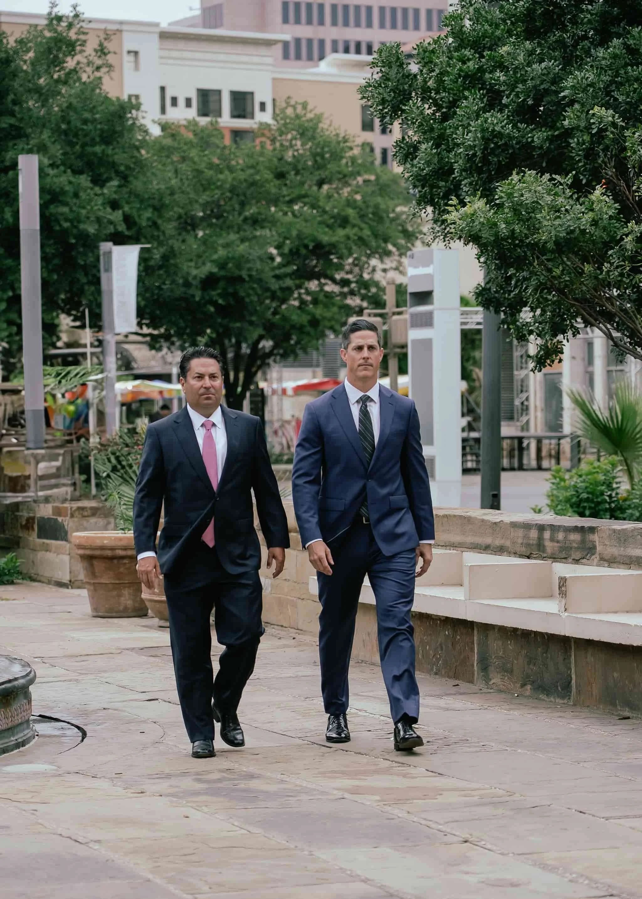 David Christian and Thom Nisbet in suits, both DWI Defense Attorney, walk side by side on a stone pathway in an urban area with trees and buildings.