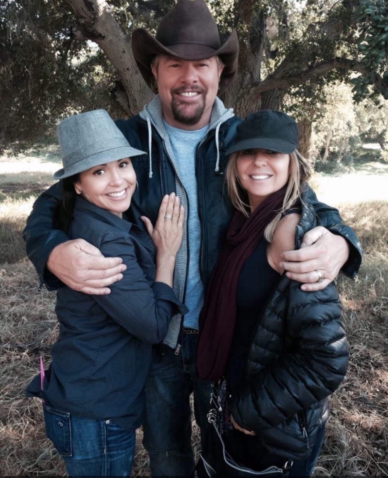 Three people outdoors hugging and smiling, two women and one man wearing jackets and hats, in front of a large tree.