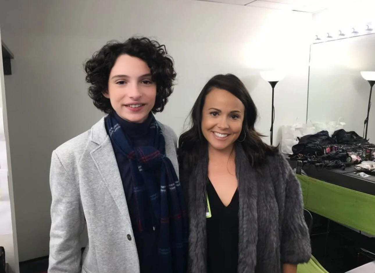 Two women smiling for a photo indoors, standing in front of a mirror with two lamps and a table with makeup and accessories.