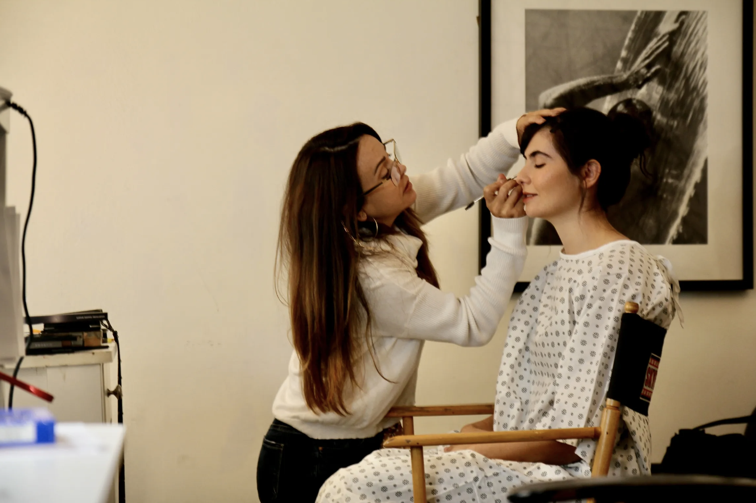 A woman with long brown hair and glasses applies makeup to another woman sitting in a chair, who has dark hair tied back and is wearing a hospital gown.