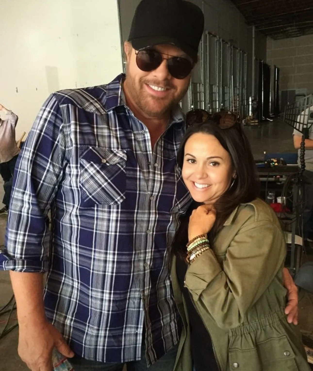 A man and a woman are smiling and posing for a photo indoors. The man is wearing a black hat, sunglasses, a blue plaid shirt, and has a beard. The woman is wearing a green jacket and has long dark hair with sunglasses resting on her head.