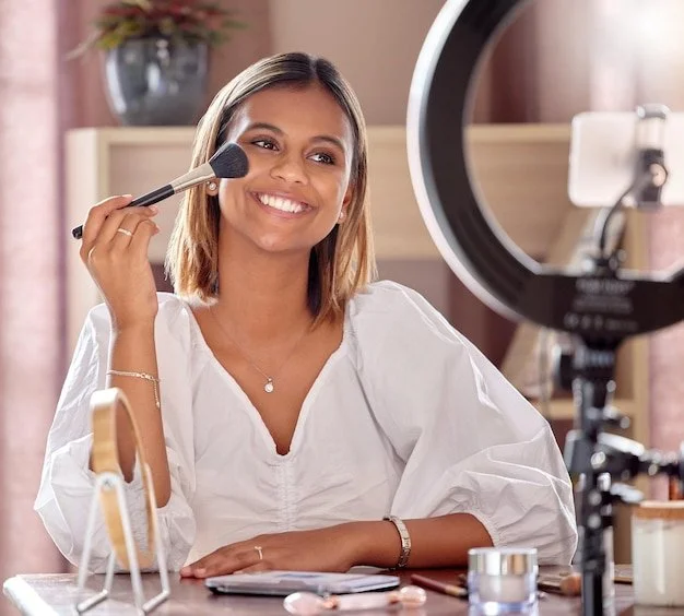 Woman applying makeup in front of a ring light, smiling, with makeup products on the table.