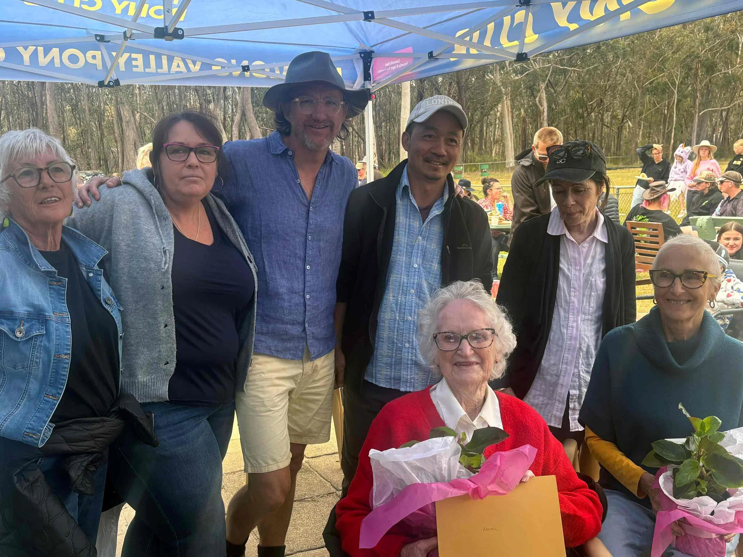 Group of eight people, including elderly woman with white hair and glasses, holding pink flowers and a plaque, gathered outdoors under a blue canopy. Other people are visible in the background at a picnic or outdoor event.