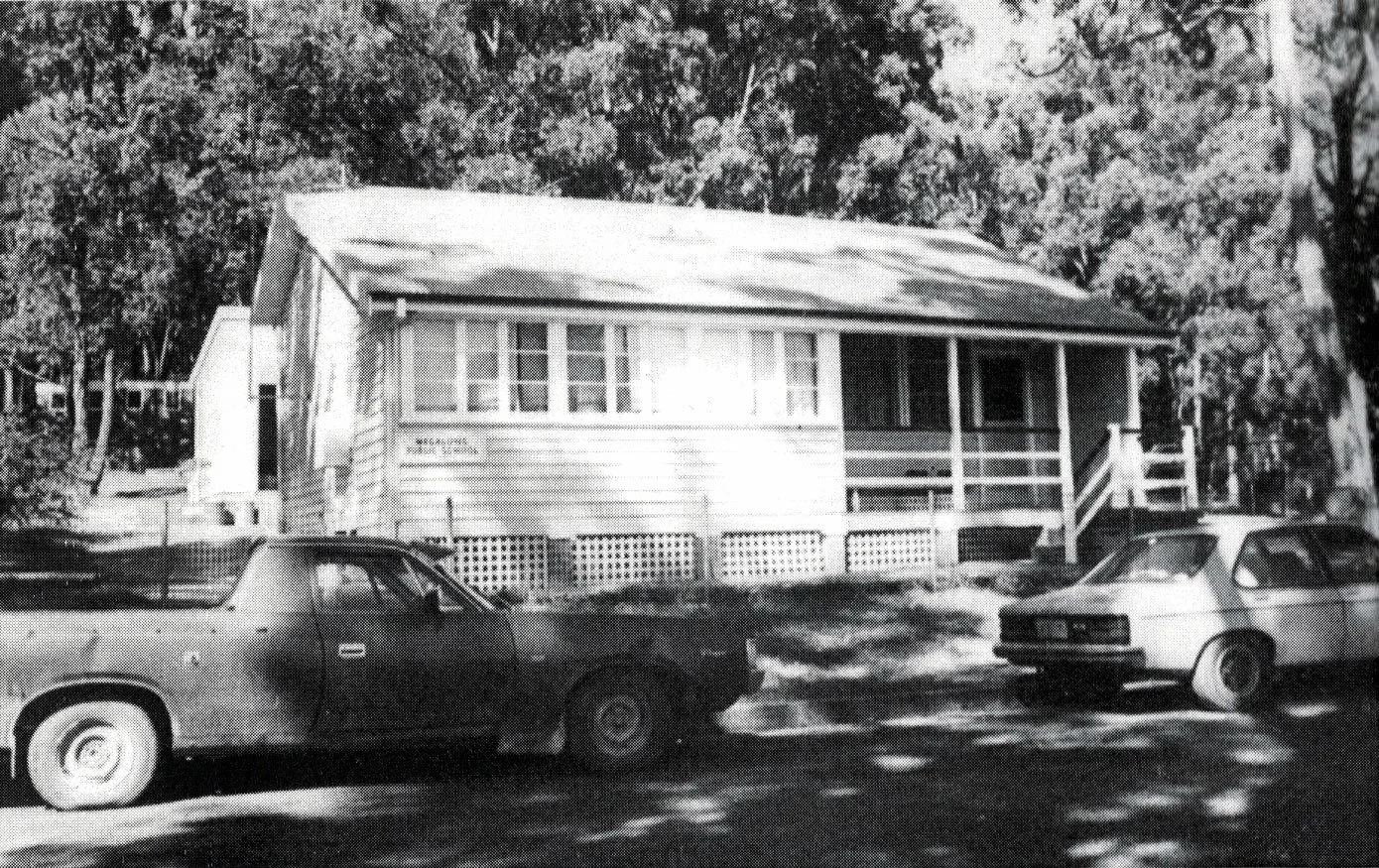 A black and white photograph of the Megalong Public School with a porch and stairs, surrounded by trees, with three cars parked in front.