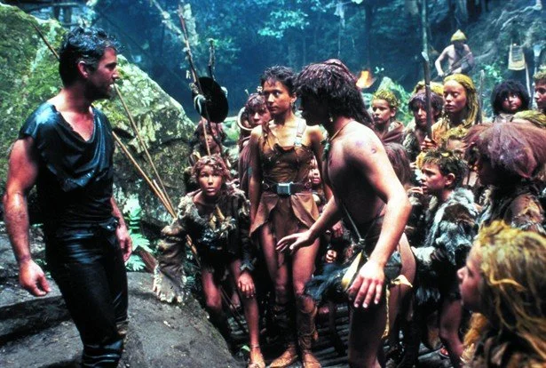 Scene from a movie set in a jungle, with actors in primitive costumes surrounded by other actors in similar attire, and a man in a black shirt.