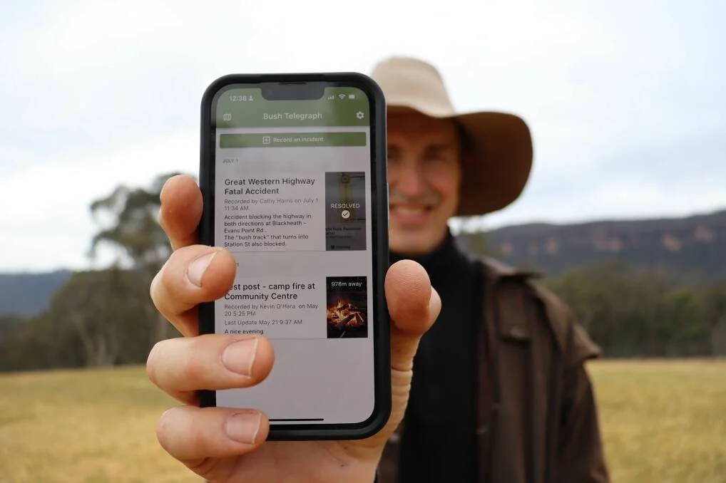 Person outdoors holding a smartphone with a news app displaying accident reports, blurred background with grass and hills.