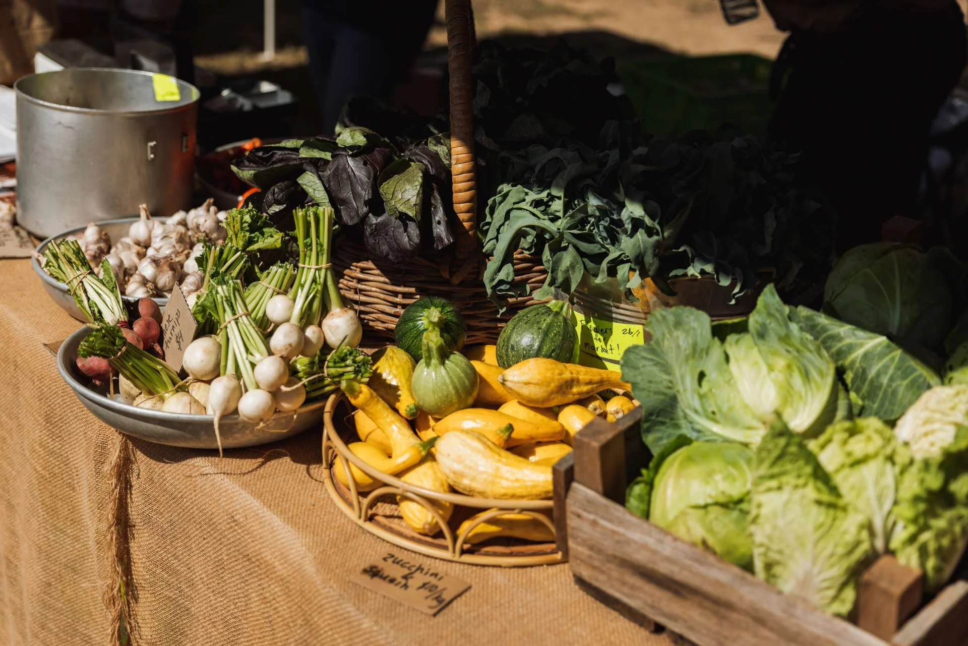 Fresh vegetables on display at a market stall, including turnips, zucchini, yellow squash, leafy greens, and lettuce in baskets and bowls with a burlap-covered table.