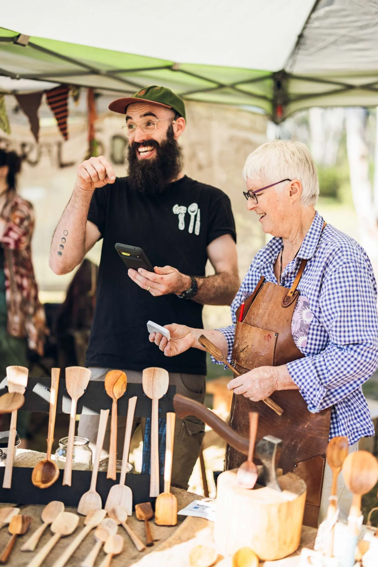 A bearded man and an older woman selling wooden kitchen utensils at an outdoor market stall under a canopy, with the man holding a smartphone and the woman wearing an apron and checking her phone, both smiling.