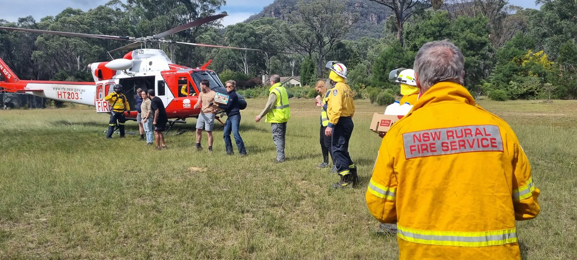 NSW Rural Fire Service personnel and civilians gather on a grassy field near a red and white rescue helicopter painted with NSW Government markings, with trees and hills in the background.