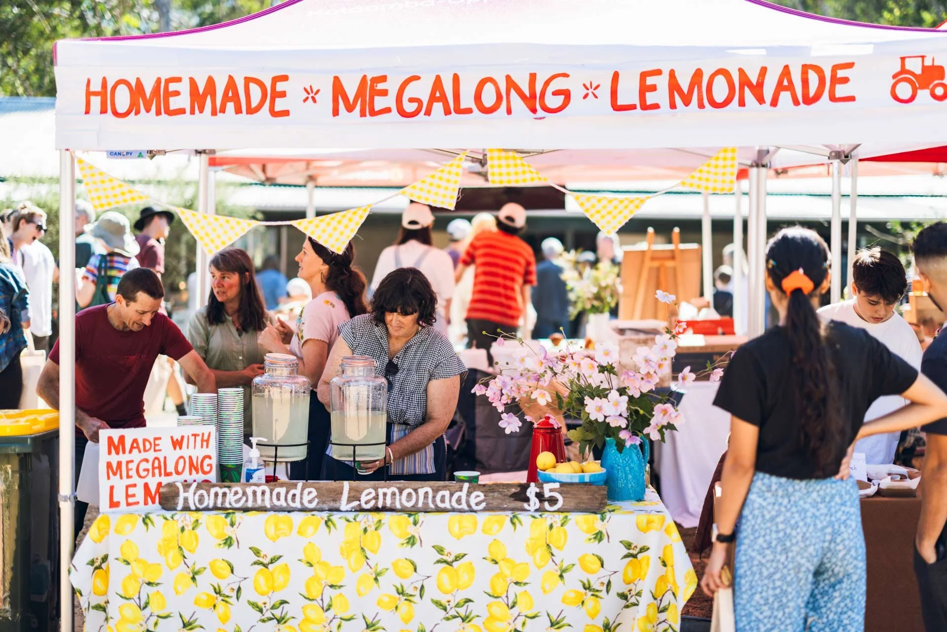 A busy outdoor market stand selling homemade megalong lemonade with a sign indicating it costs five dollars, decorated with a lemon-themed tablecloth and flowers, with several people browsing and serving customers.