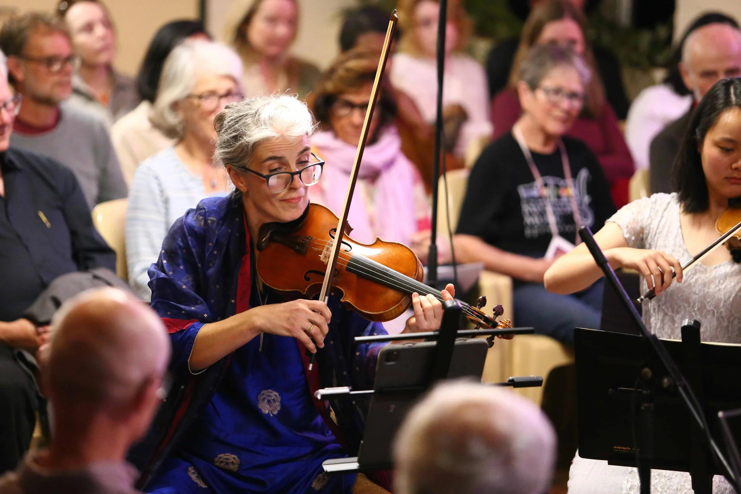 A woman with white hair and glasses playing a violin in a concert hall, surrounded by an audience.