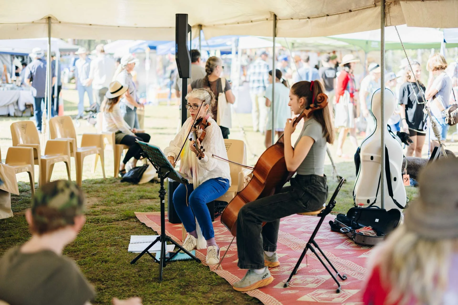 Two women playing string instruments, a violin and a cello, under a tent at an outdoor event, with a crowd of people browsing vendor booths in the background.