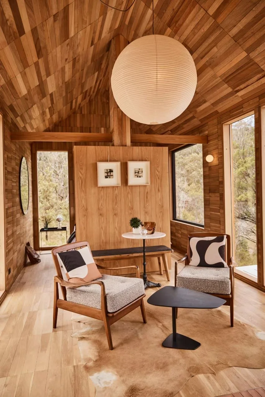 A cozy living room with wood-paneled vaulted ceiling and walls, large windows showing trees outside, a large round paper lantern hanging from the ceiling, a small round white table with decorative items, two mid-century modern armchairs with patterned cushions, a cowhide rug, and framed artwork on a wooden screen.
