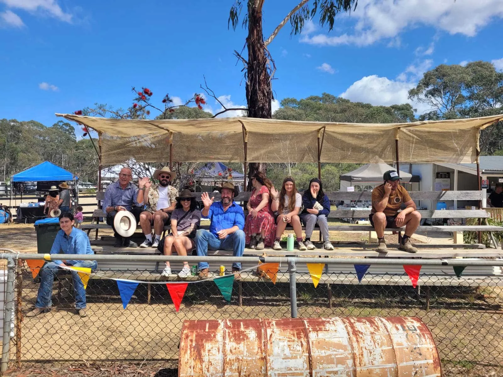 Group of ten people sitting on spectator benches in an outdoor setting with trees and blue sky in the background. Two people are waving and one is talking on a phone, with colorful bunting hanging in front.