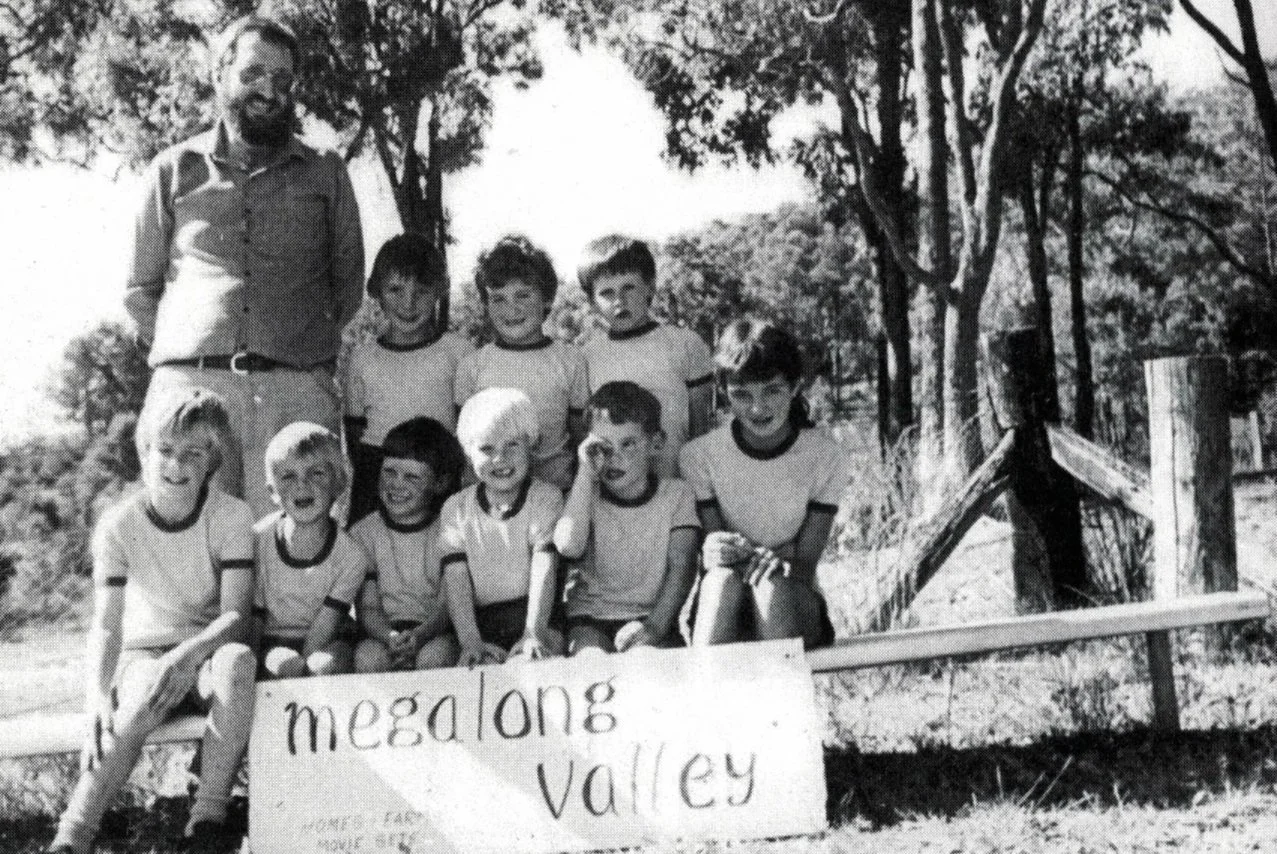 Black and white photo of a group of school children and their teacher posing outdoors behind a sign that reads 'Megalong Valley.' The children are sitting and standing in front of a wooden fence and trees.