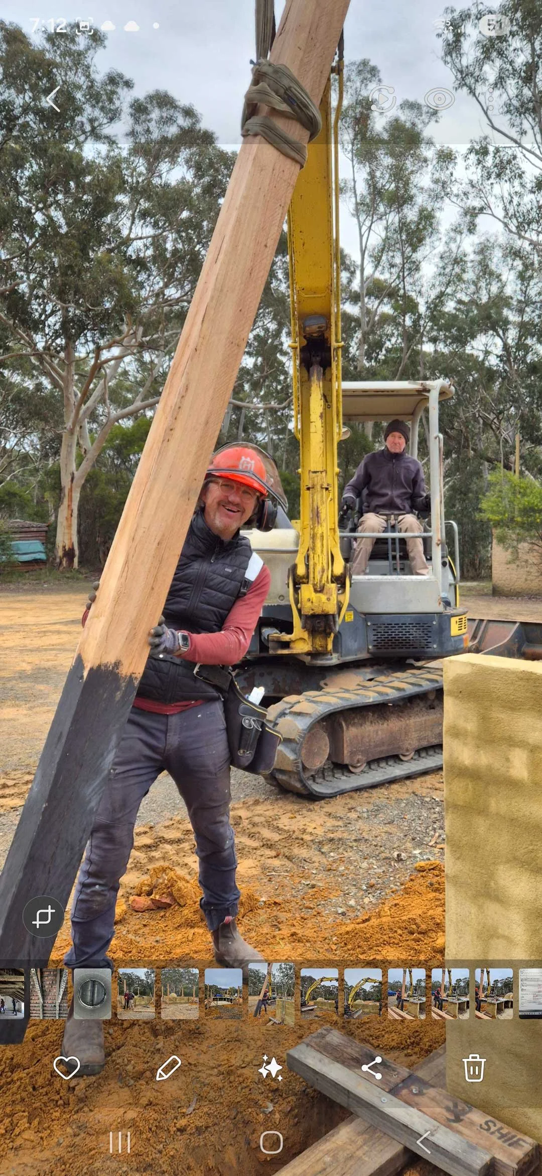 Construction site with two workers and a small excavator, one worker smiling and wearing a helmet, the other sitting on the excavator, surrounded by trees and construction materials.