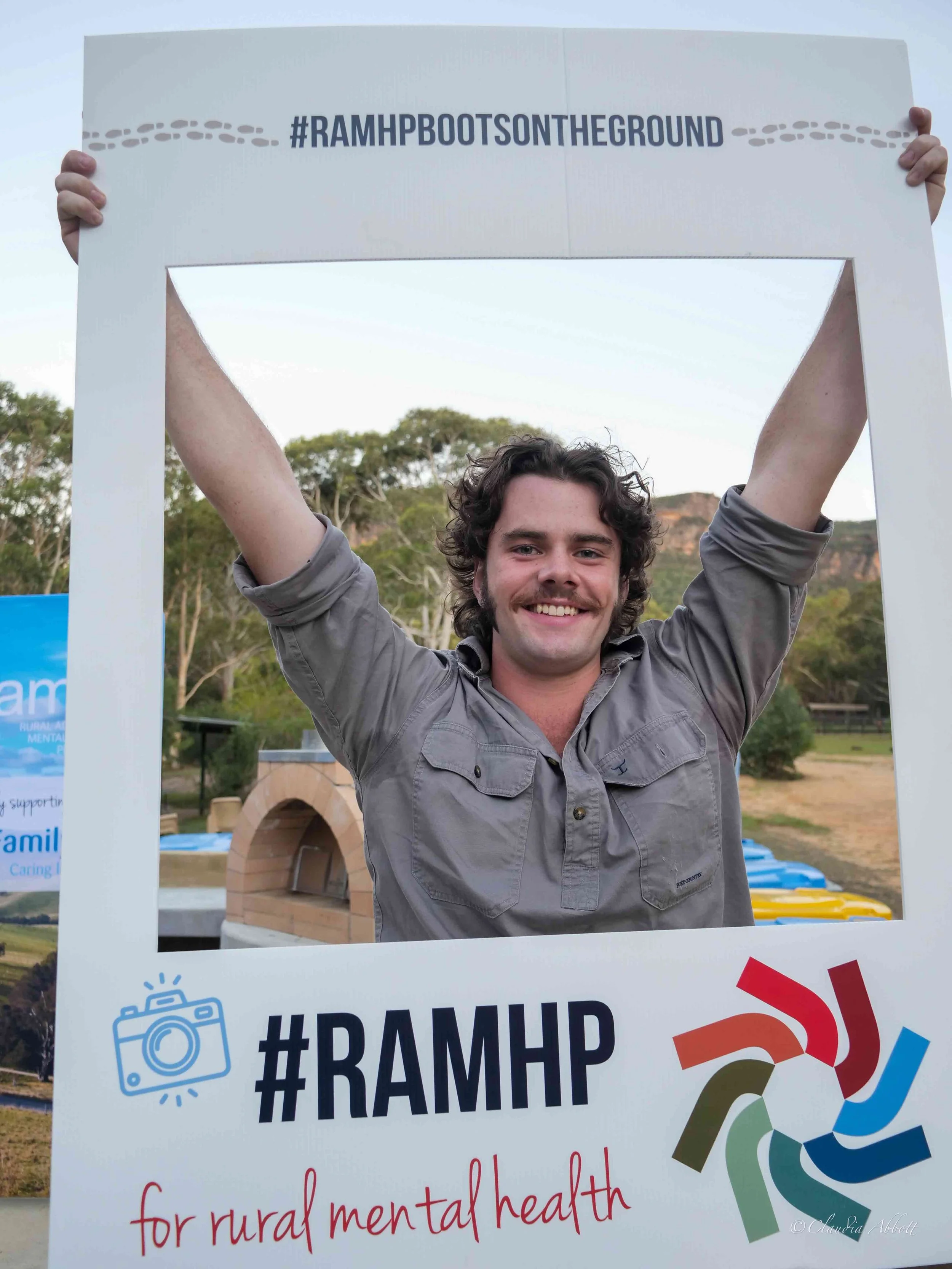 Young man smiling and holding a large white frame with hashtags #RAMHP and text supporting rural mental health, outdoors with trees and a clay oven in the background.