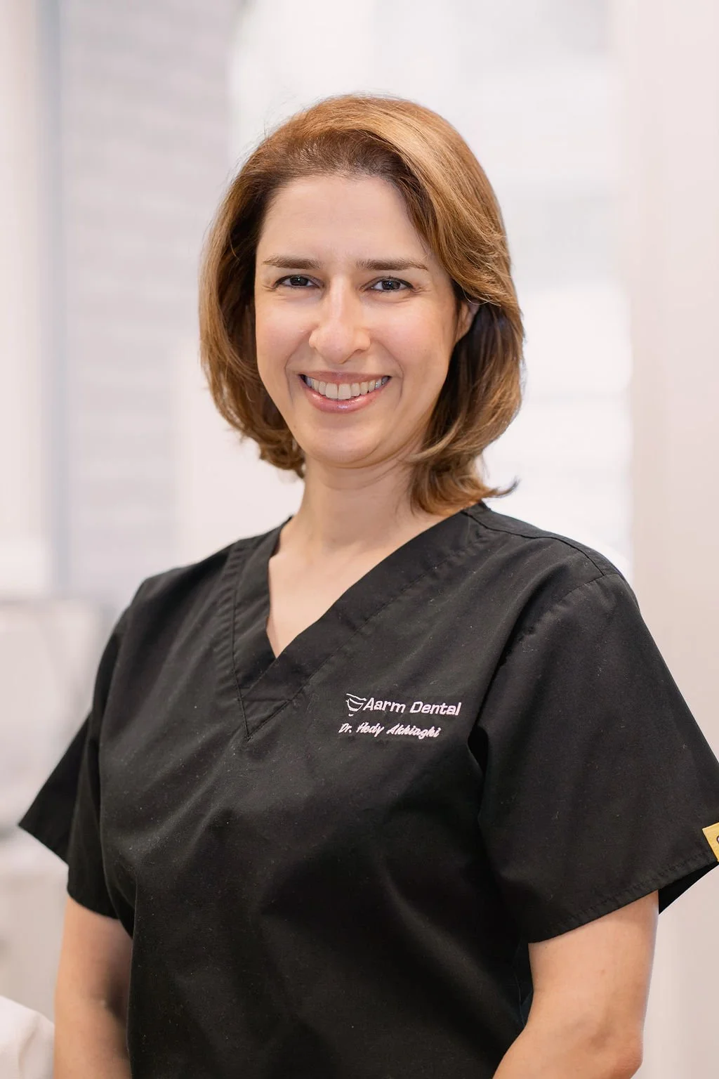 A smiling woman wearing a black dental uniform with the logo 'Arm Dental' and the name 'Dr. Hedy Almazoglu' embroidered on it, standing in what appears to be a dental office.