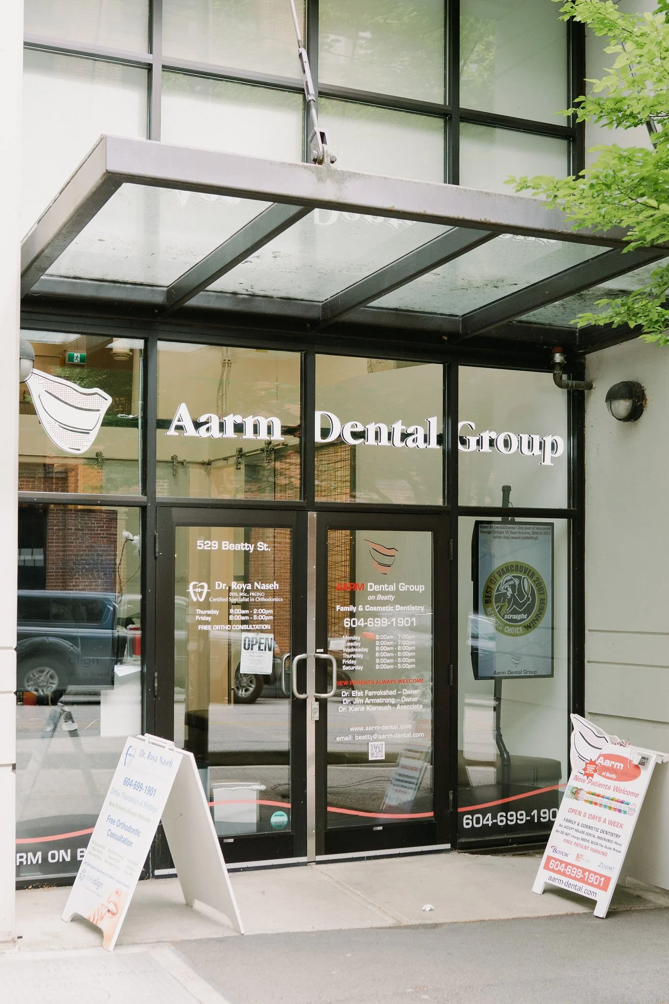 The entrance of Aarm Dental Group with glass doors and windows, displaying the clinic's name and contact information, along with signs advertising services and hours.