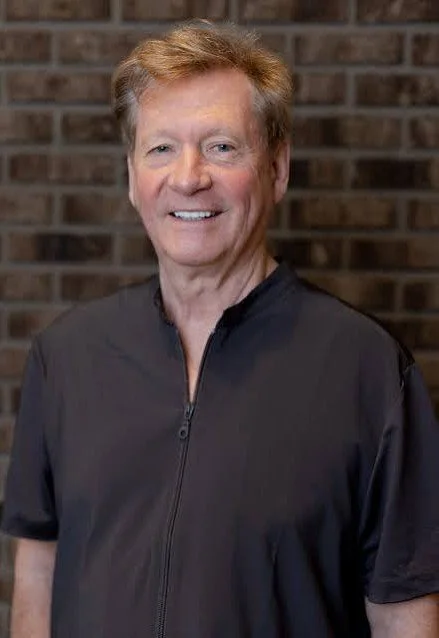 A smiling man with light brown hair, wearing a black short-sleeved zip-up shirt, standing in front of a brick wall.