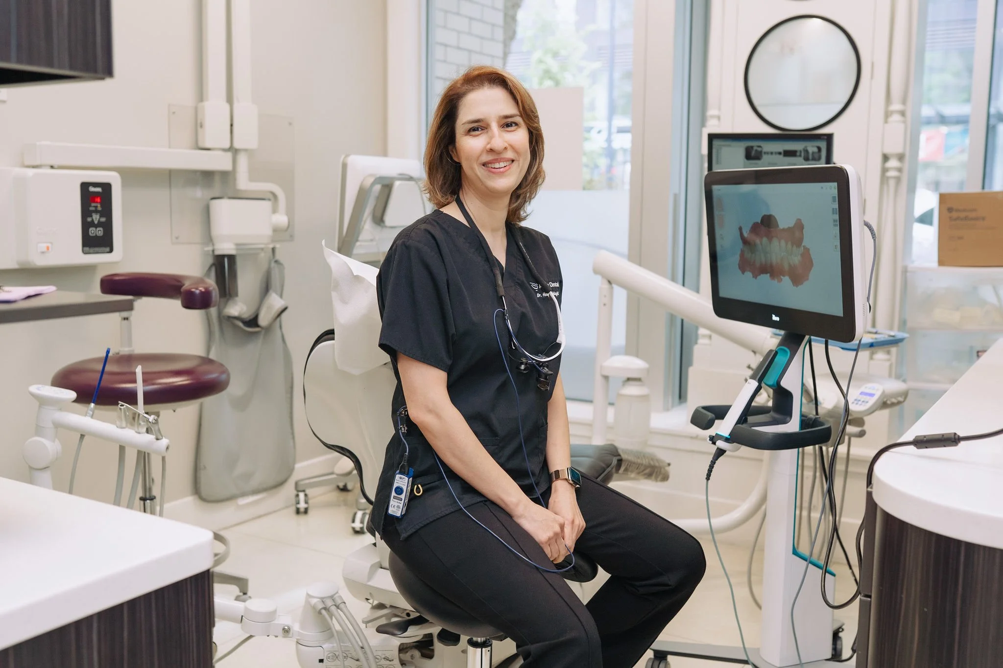 A woman in dental scrubs sitting in a dental office with a monitor showing a digital scan of teeth.
