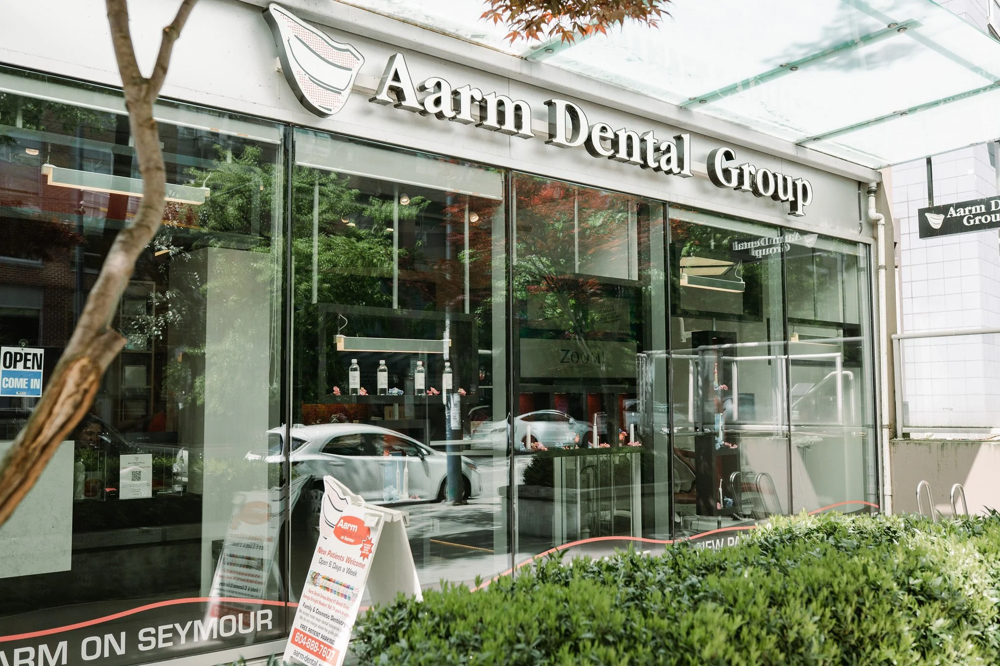 The exterior of an Aarm Dental Group office with glass windows reflecting the street and cars. Signage is visible with the office name and logo. A promotional signboard stands outside near greenery.