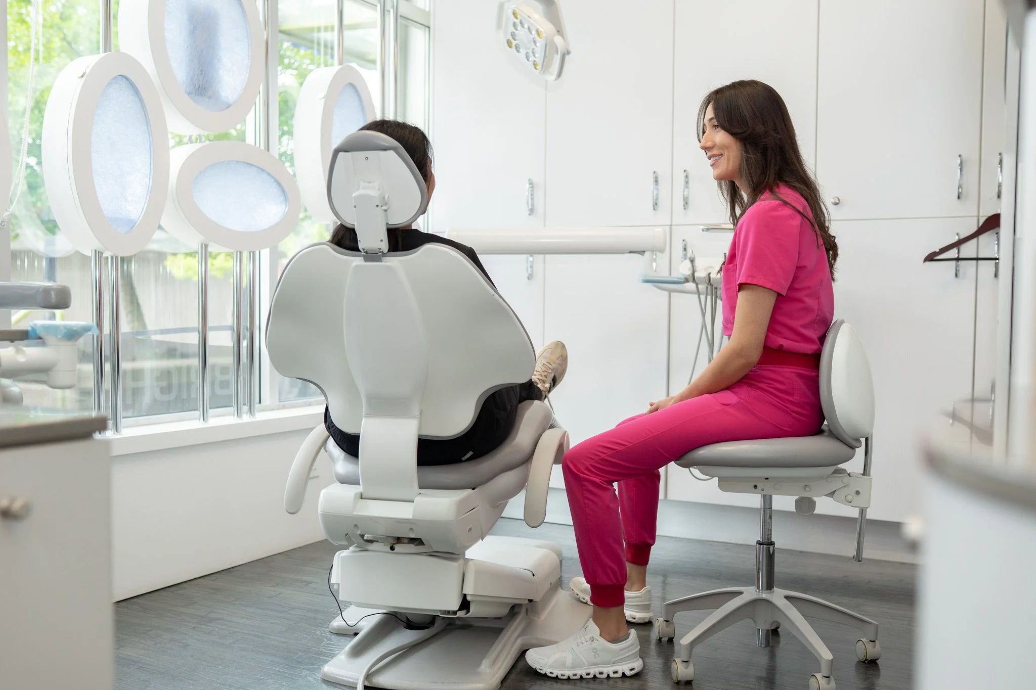 Dentist or dental professional talking to a patient sitting in a dental chair in a dental clinic with large windows.
