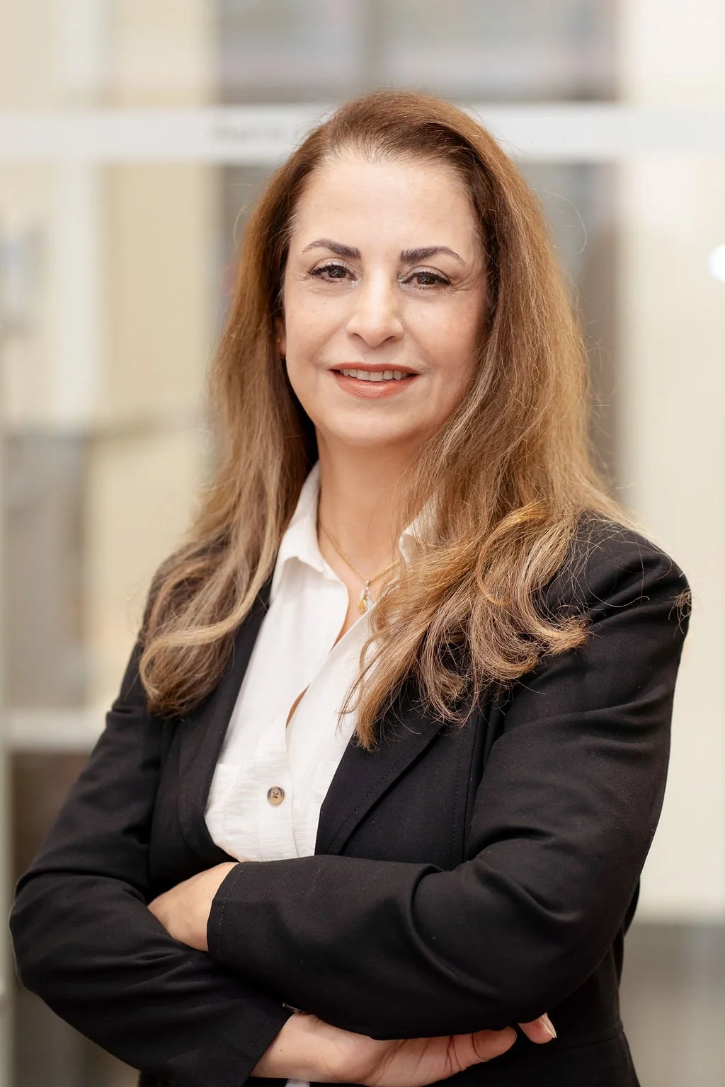 A professional woman with long, wavy hair, smiling, wearing a black blazer and white shirt, standing with arms crossed in an indoor office setting.