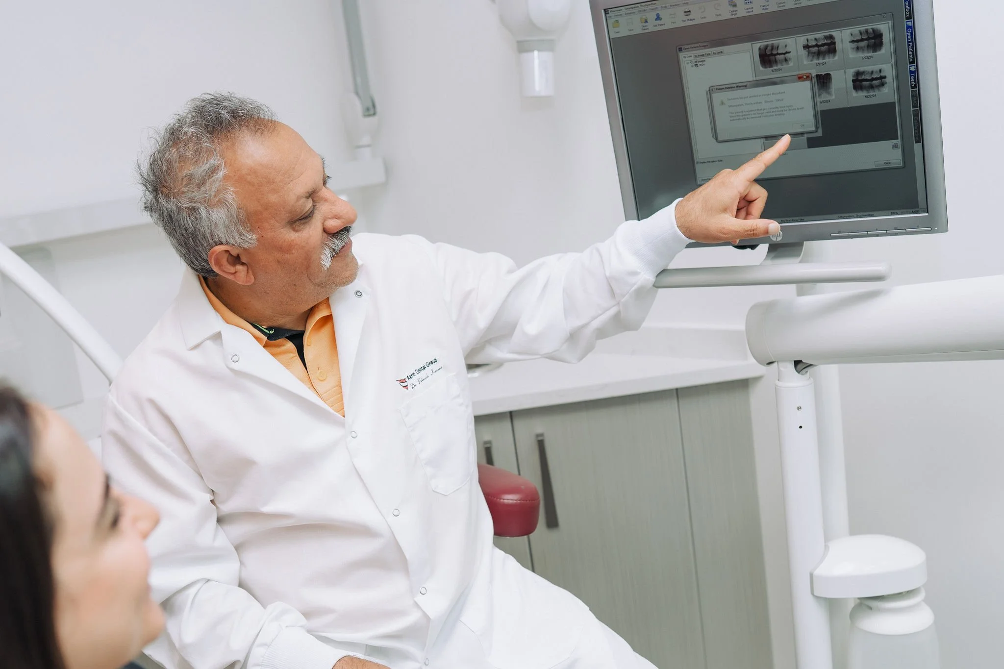 A doctor in a white coat points at a monitor displaying dental X-ray images in a medical office.