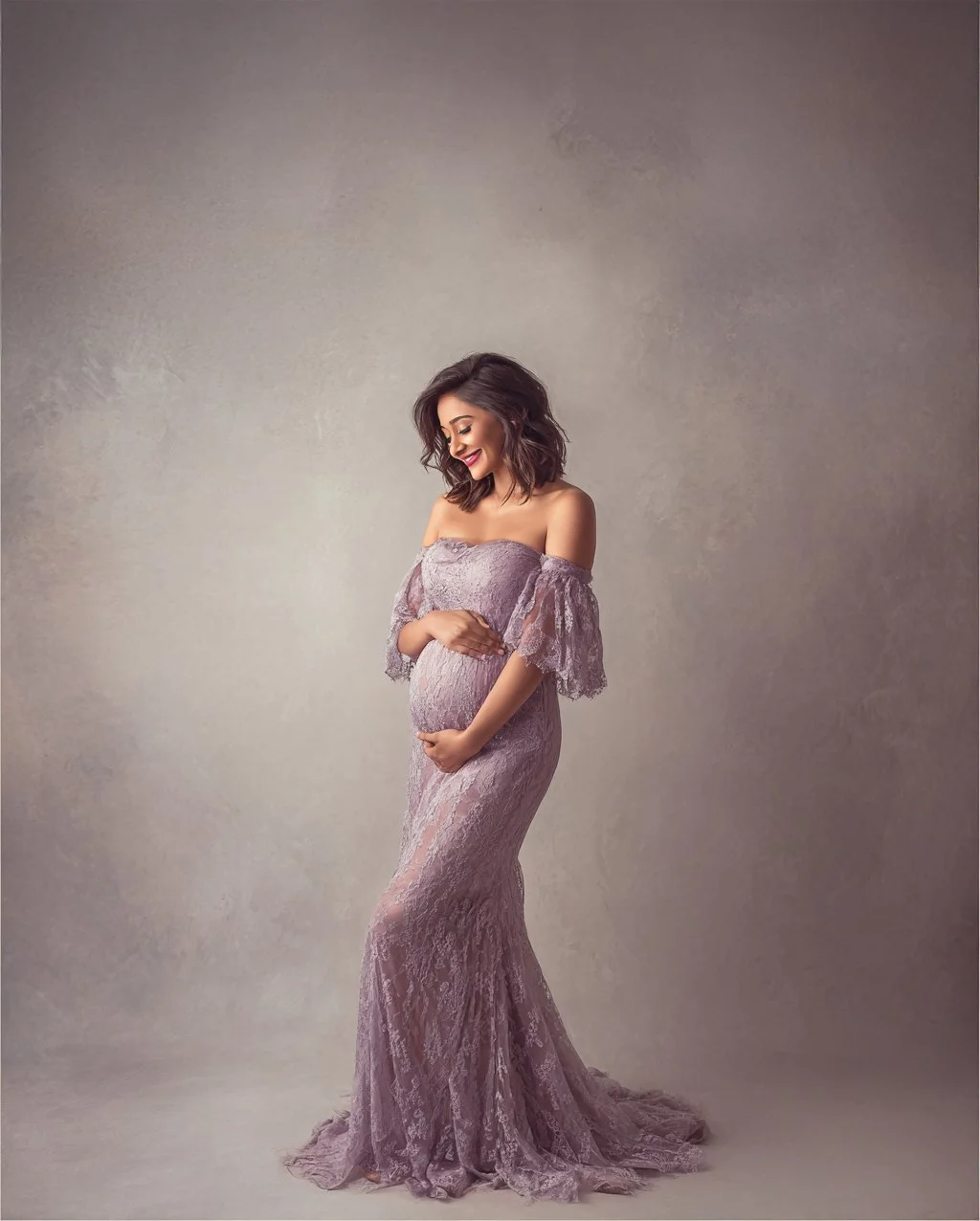 Pregnant woman in a lavender lace off-shoulder gown, smiling and holding her belly, standing against a neutral background.