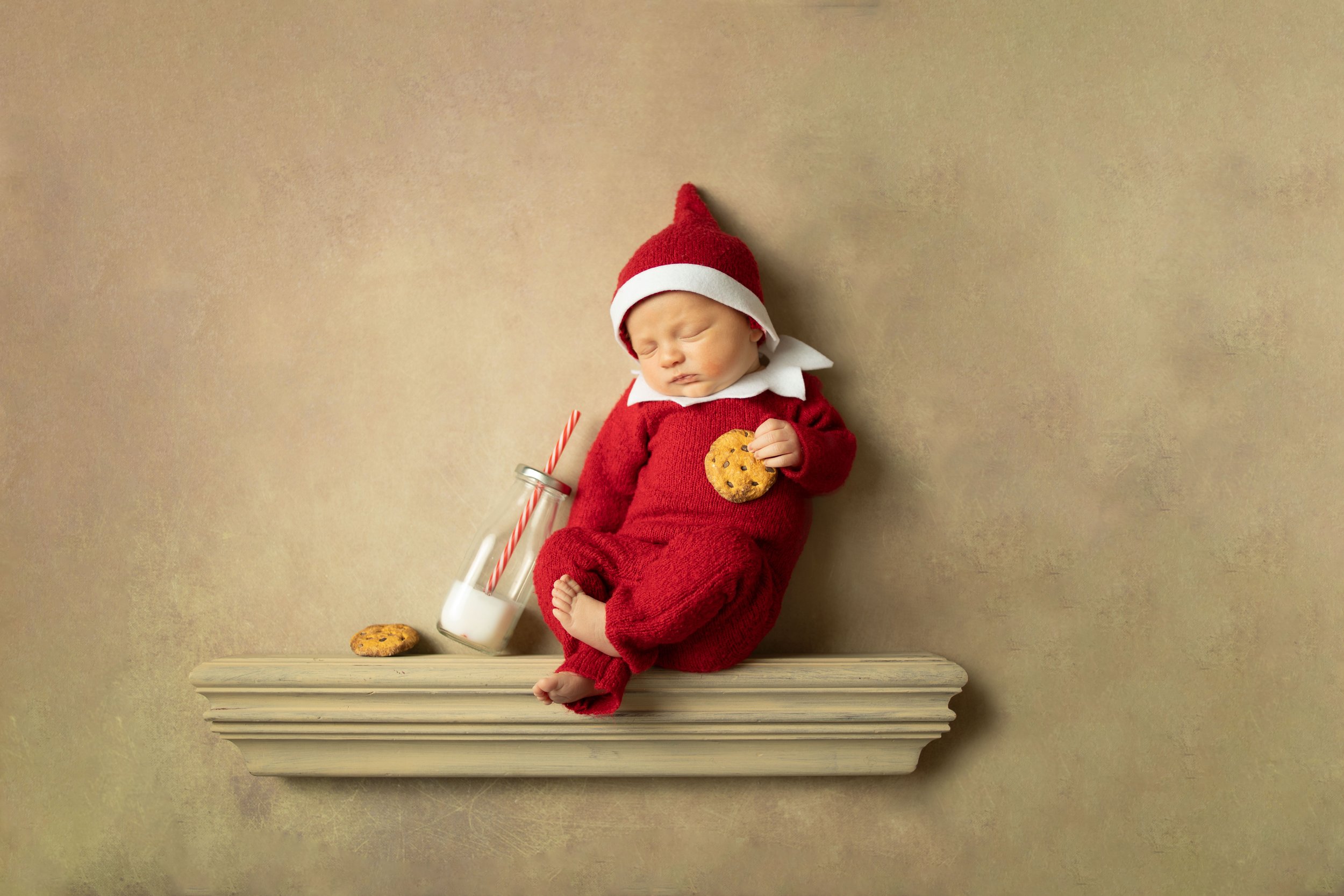 Sleeping baby in red elf costume holding a cookie, with a bottle of milk and cookie nearby, set on a shelf.