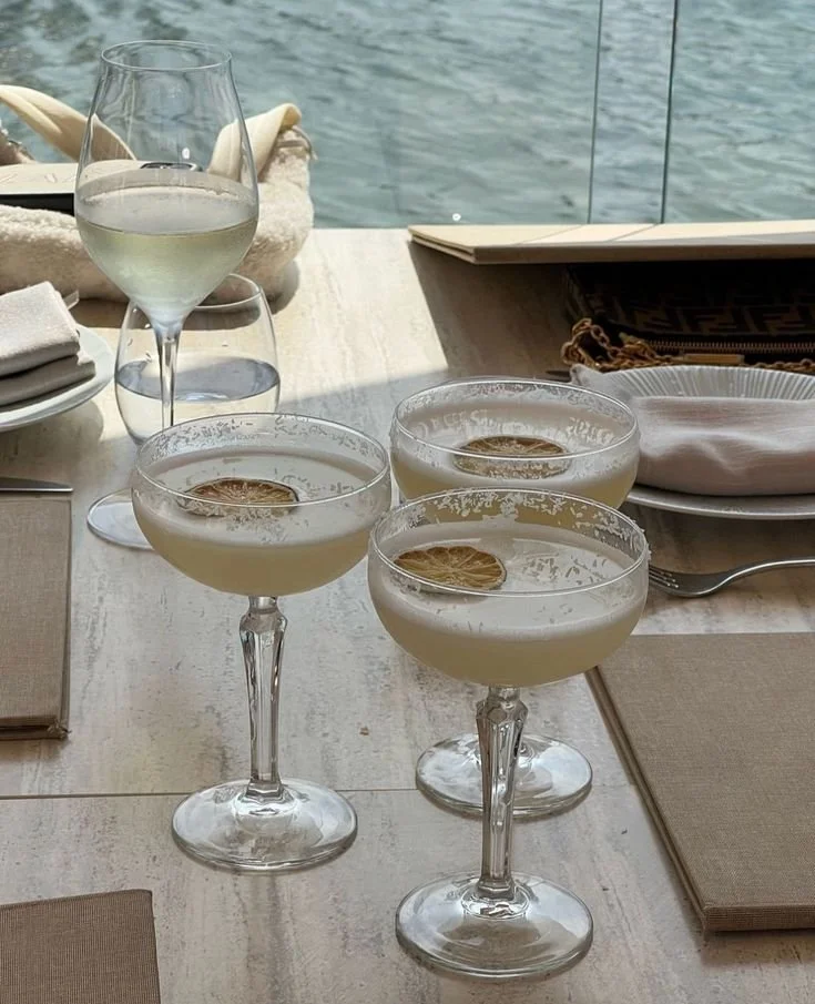 Table setting with three glasses of cocktails garnished with dried citrus slices, a glass of white wine, napkins, and a body of water in the background.