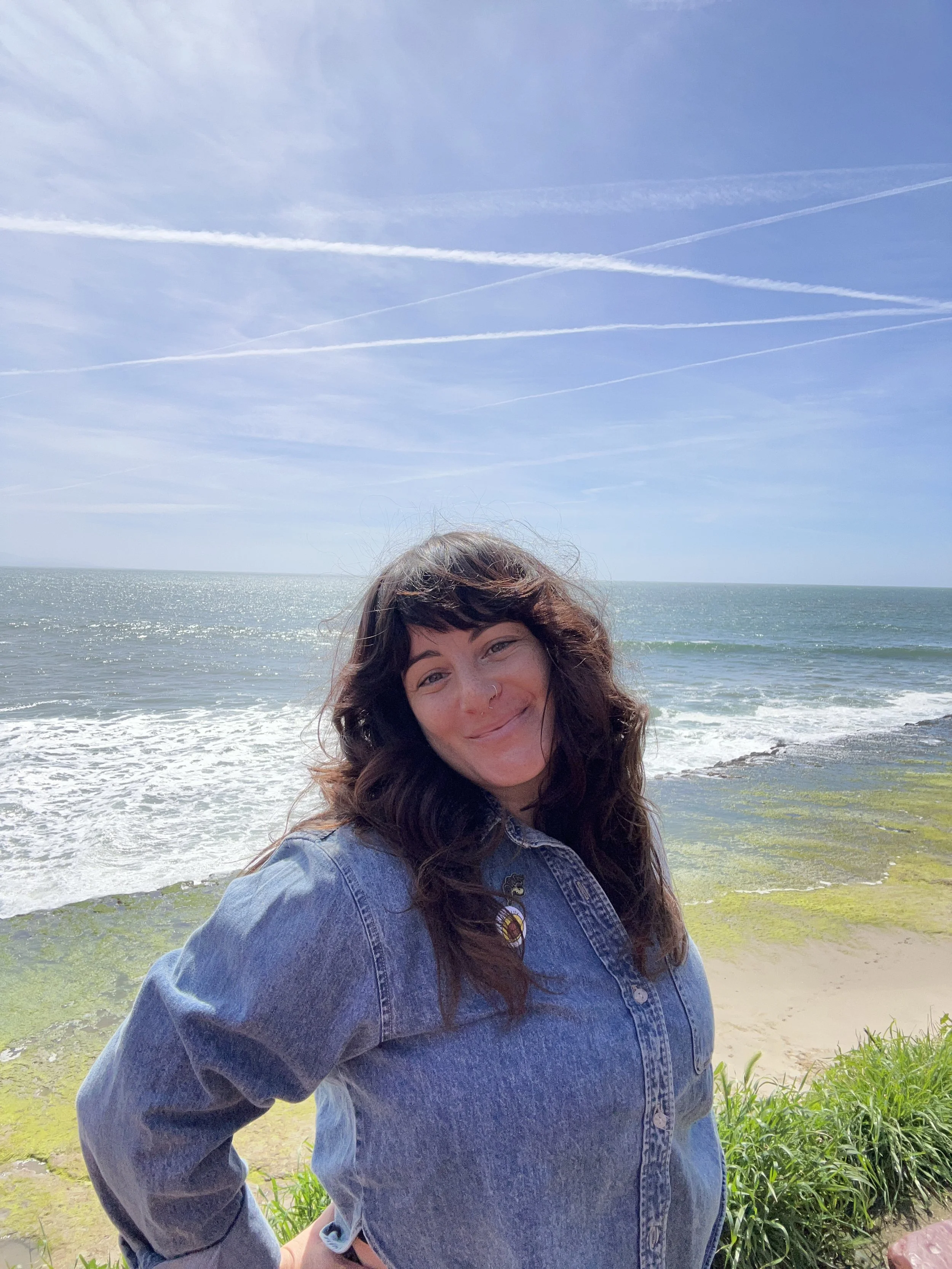 A smiling woman with dark curly hair wearing a denim jacket stands on a beach with waves, sandy shore, and blue sky with contrails in the background.