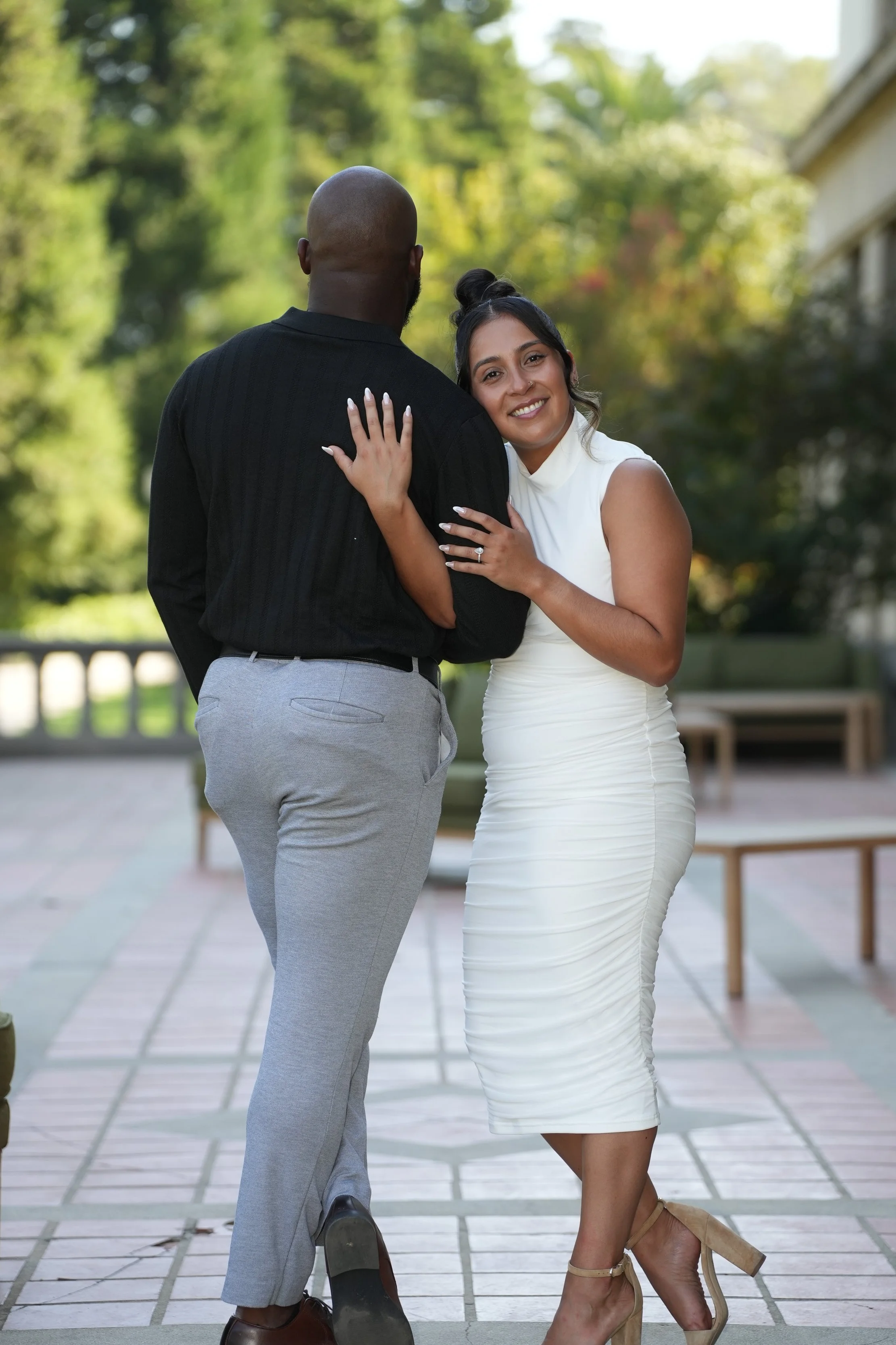 A woman in a white dress smiling while embracing a man in a black shirt and gray pants, with trees in the background.