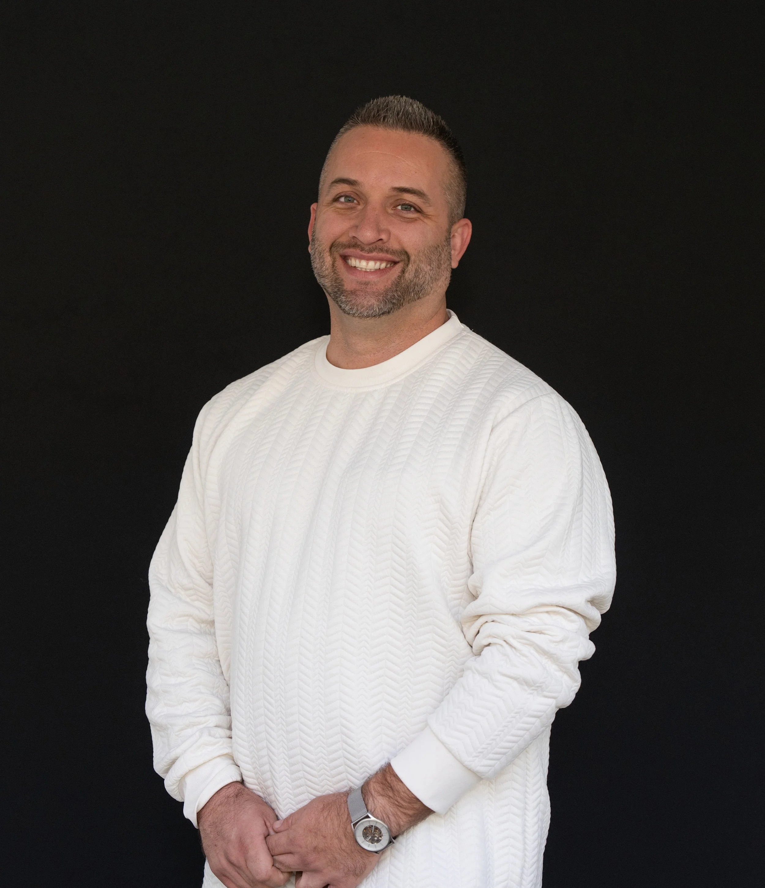 Professional headshot man in white sweater smiling with black backdrop
