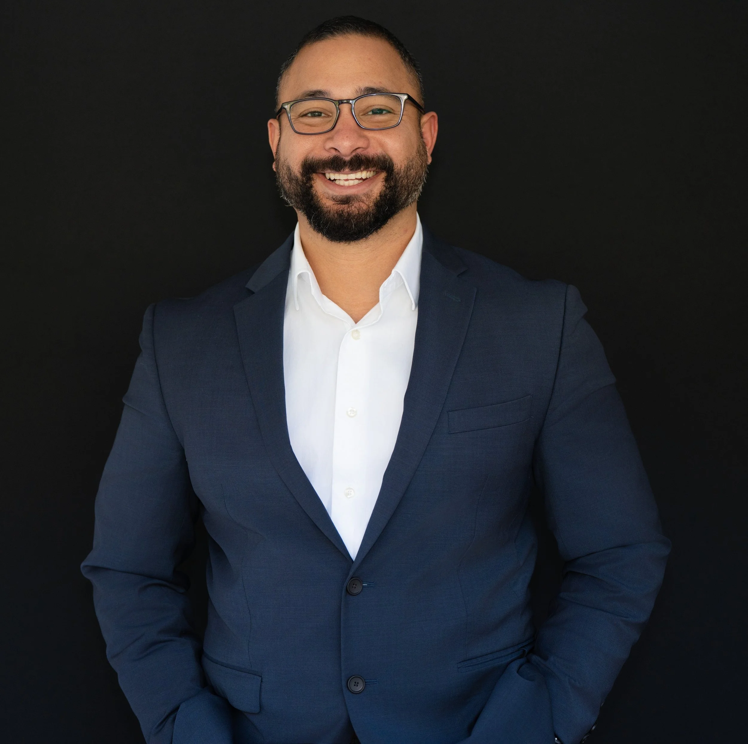 Professional headshot men in blue suit with glasses black background white shirt
