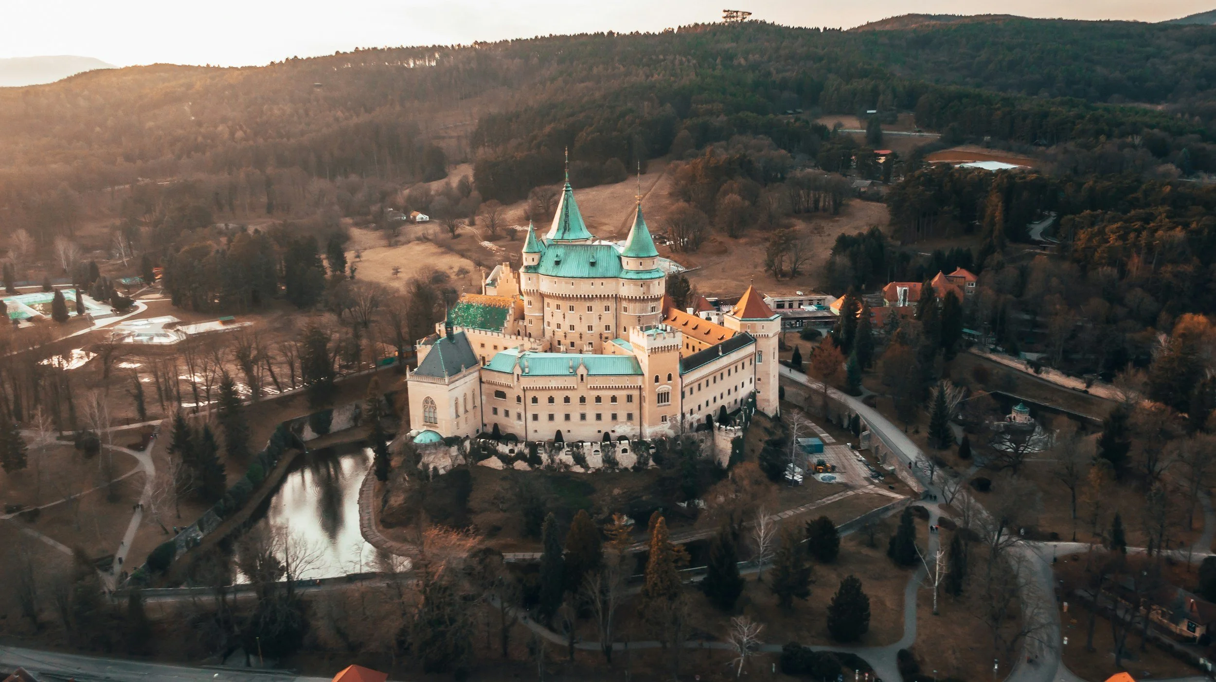 Aerial view of a castle with a river surrounding it, situated in a hilly, wooded landscape during late afternoon.