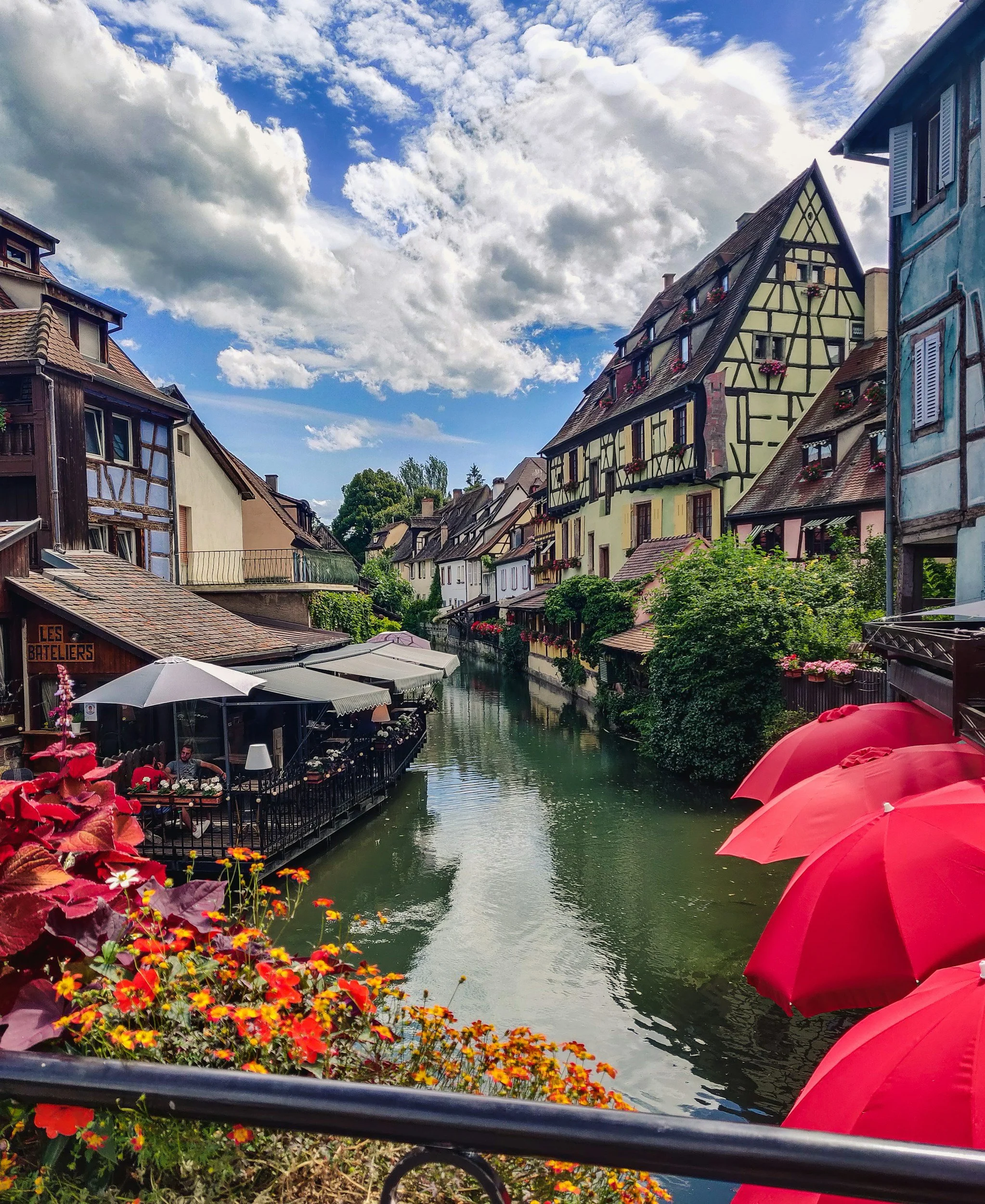 A picturesque European town with colorful, half-timbered houses lining a small canal, outdoor cafes with umbrellas, and a cloudy sky overhead.