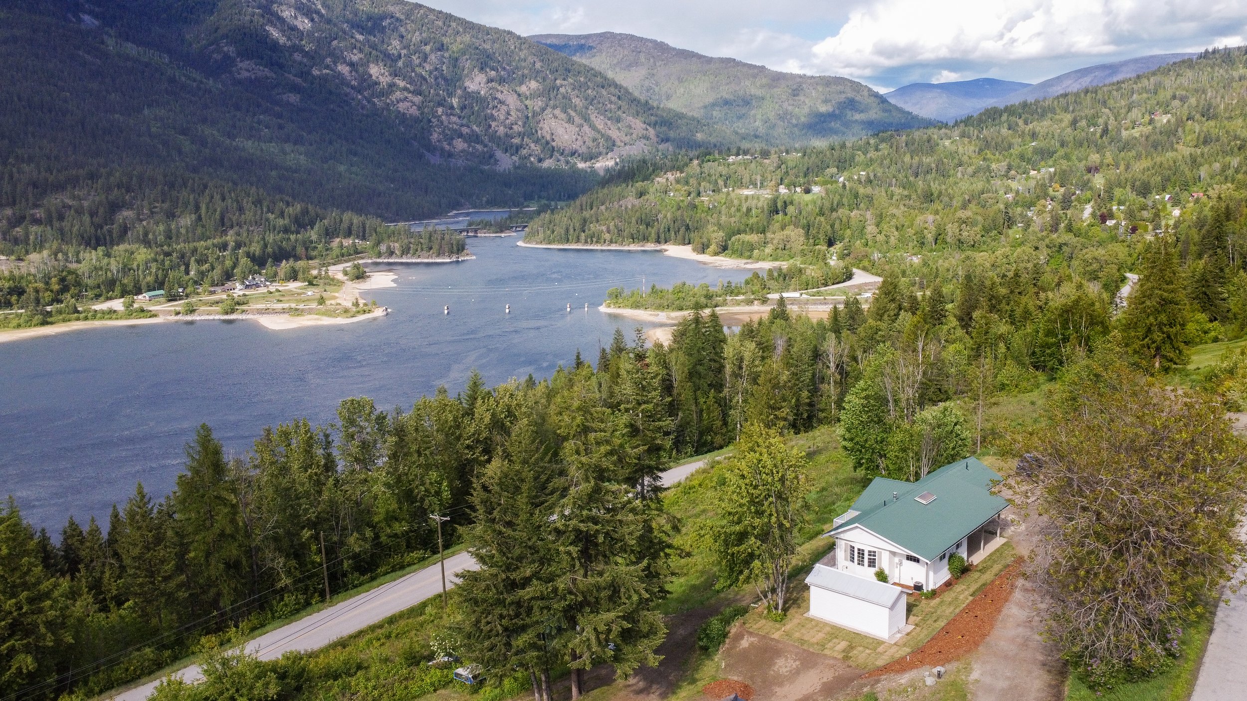 A scenic aerial view of a house with a green roof nestled among trees near a winding river, with forested hills and mountains in the background under a partly cloudy sky.