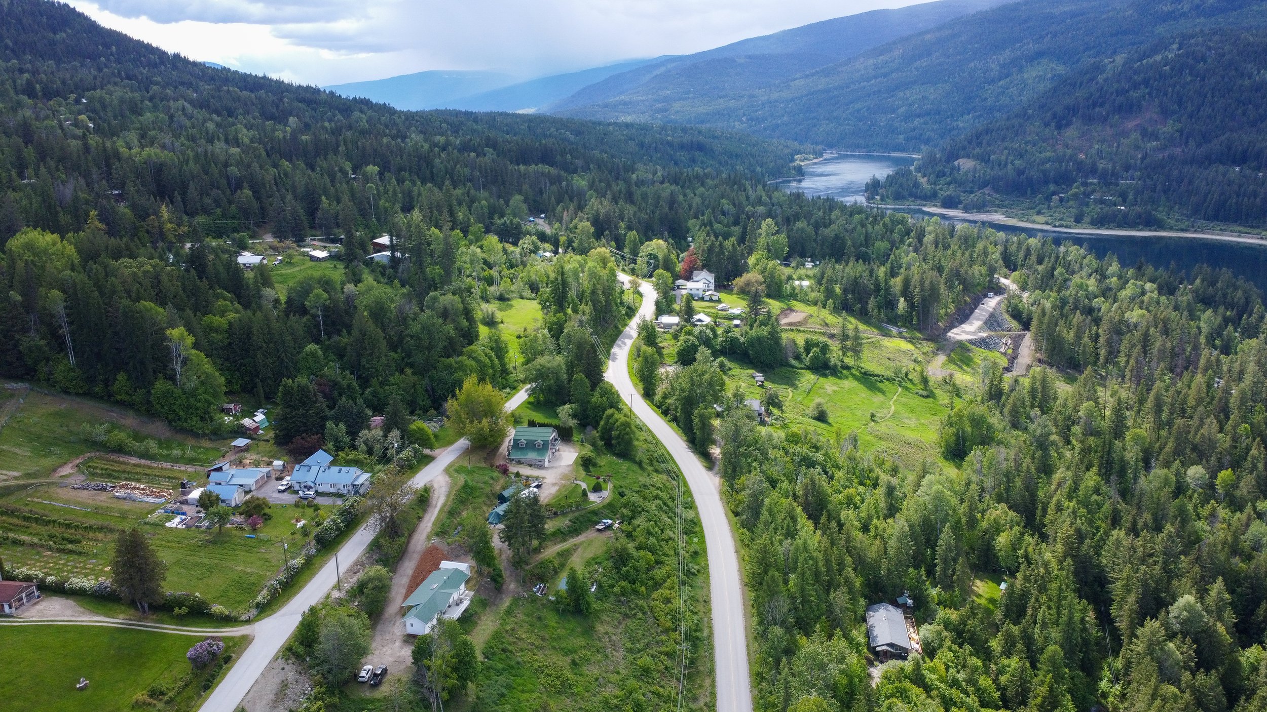 Aerial view of a green, mountainous landscape with a winding road passing through a small residential area, surrounded by dense forests and a body of water in the distance, under a cloudy sky.