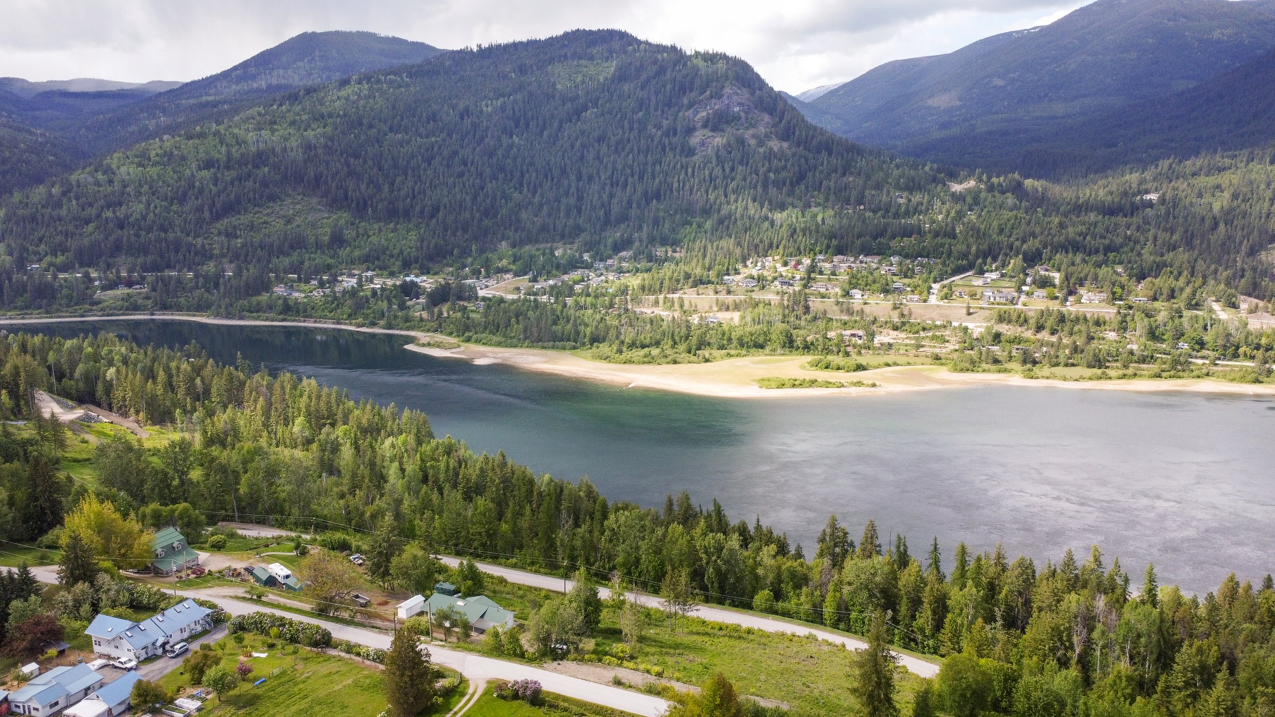Aerial view of a lake surrounded by green forests, hills, and a small residential area with houses and roads in a mountainous landscape.