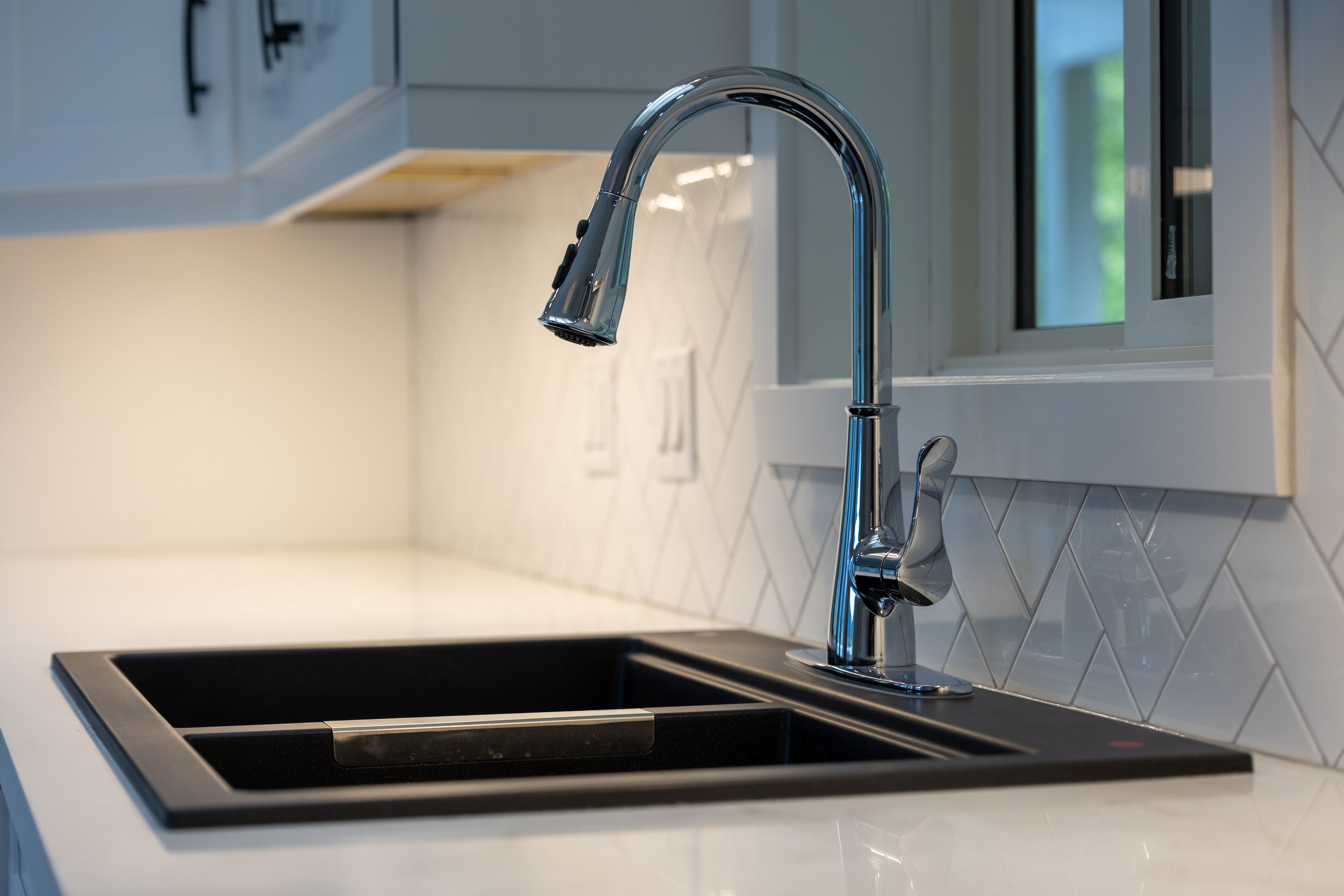 Modern kitchen sink with a chrome faucet, white tiled backsplash, and a window in the background.